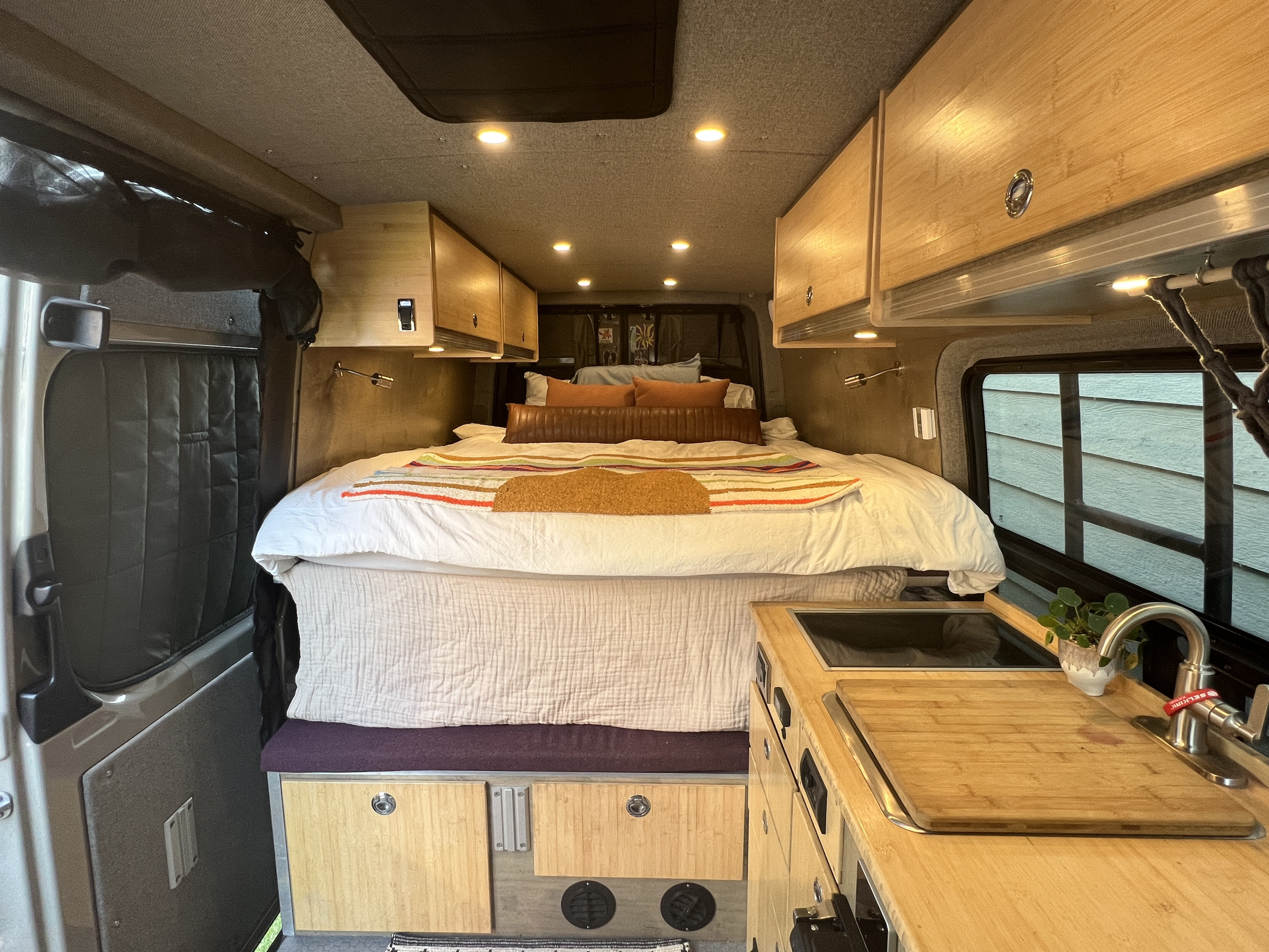 Inside a modern RV rental: a spacious bed, overhead storage, and kitchen area with a sink & cutting board.. Mercedes Benz Sprinter Van 2017