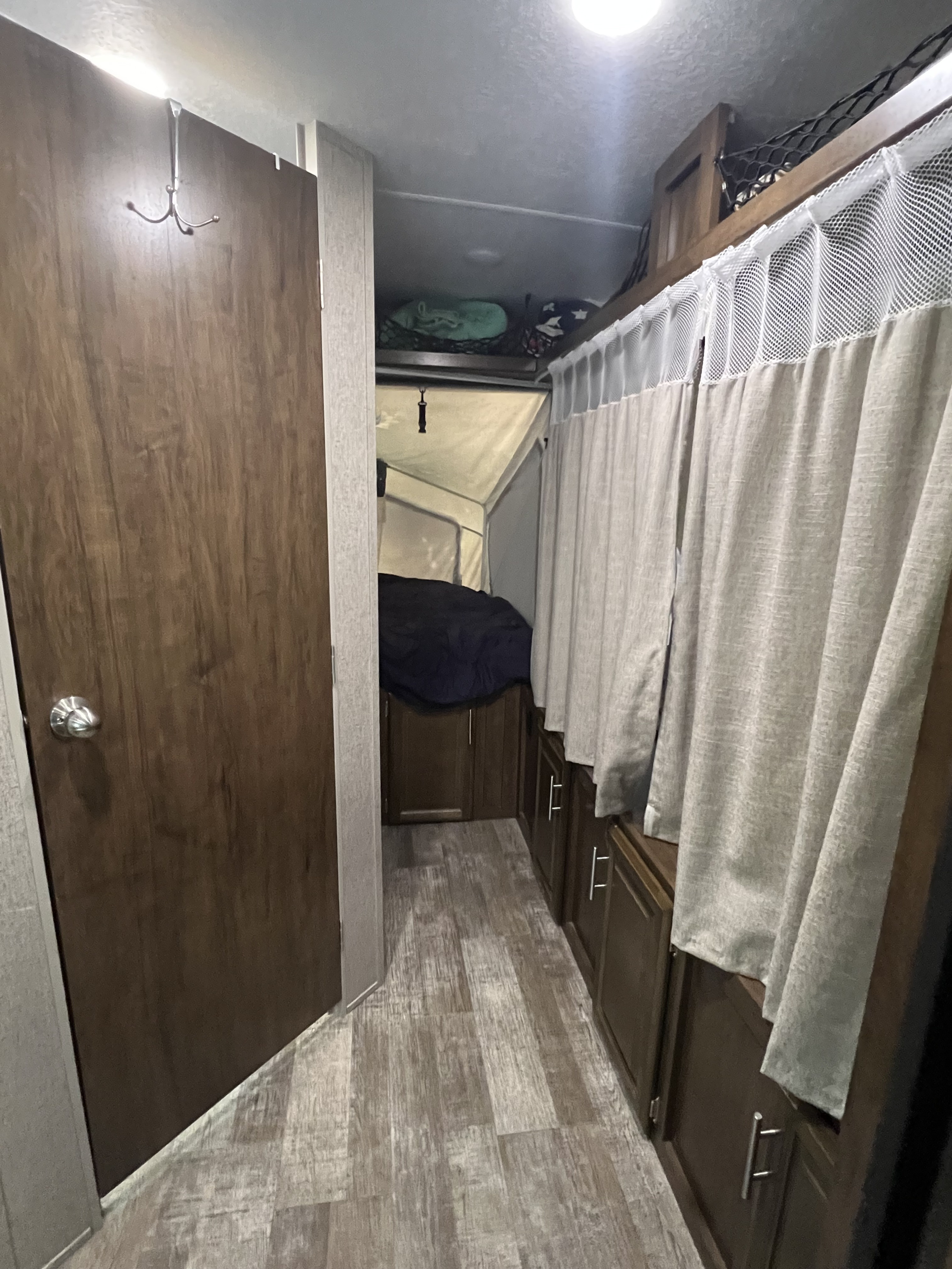 Inside an RV rental, a hallway features a wooden door, laminate flooring, and cabinets with curtains, leading to a pop-up bed area.. Forest River Rockwood Roo 2019