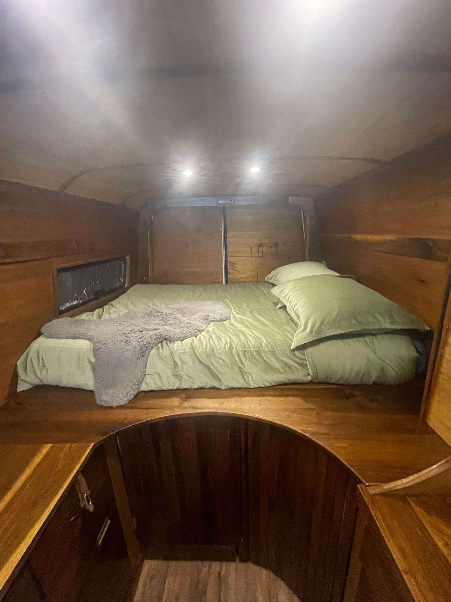 Cozy wood-paneled RV rental interior, featuring a comfy bed with green bedding and overhead lights.. Mercedes-Benz Sprinter 2019