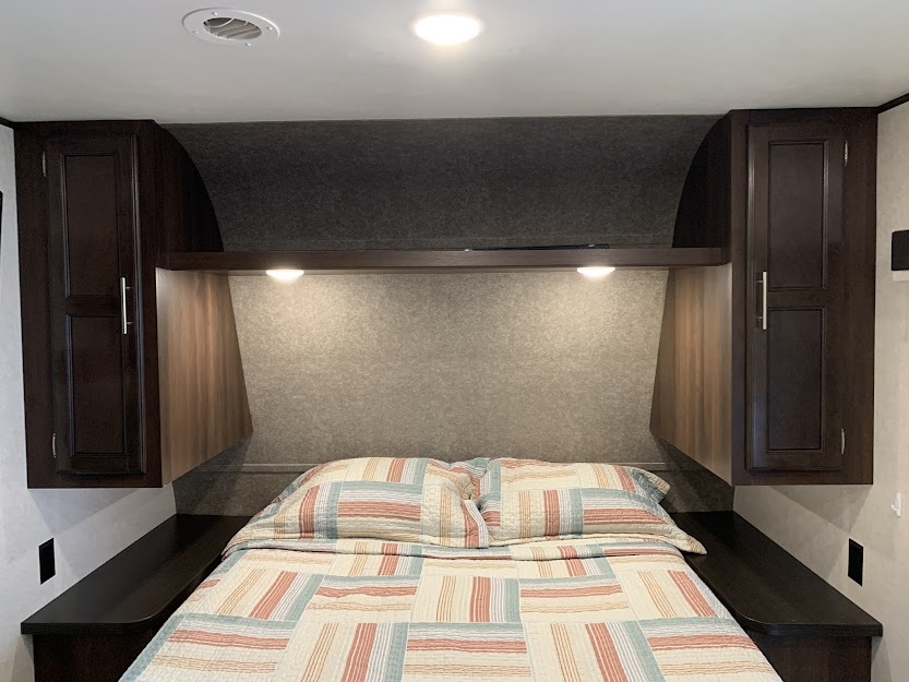 Inside an RV rental bedroom: a bed with a colorful quilt, surrounded by dark wood cabinets, shelving, and practical overhead lighting.. Jayco Jay Flight SLX 2019