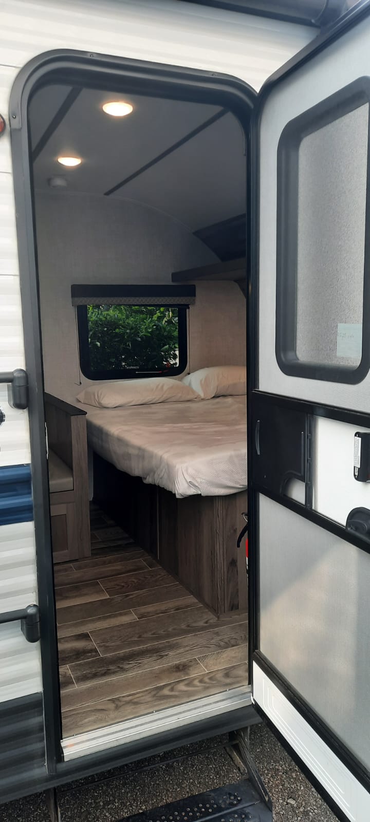 Open door reveals an inviting RV rental interior: cozy bed, window with greenery, and wood-look floor.. Grand River 19BH 2022