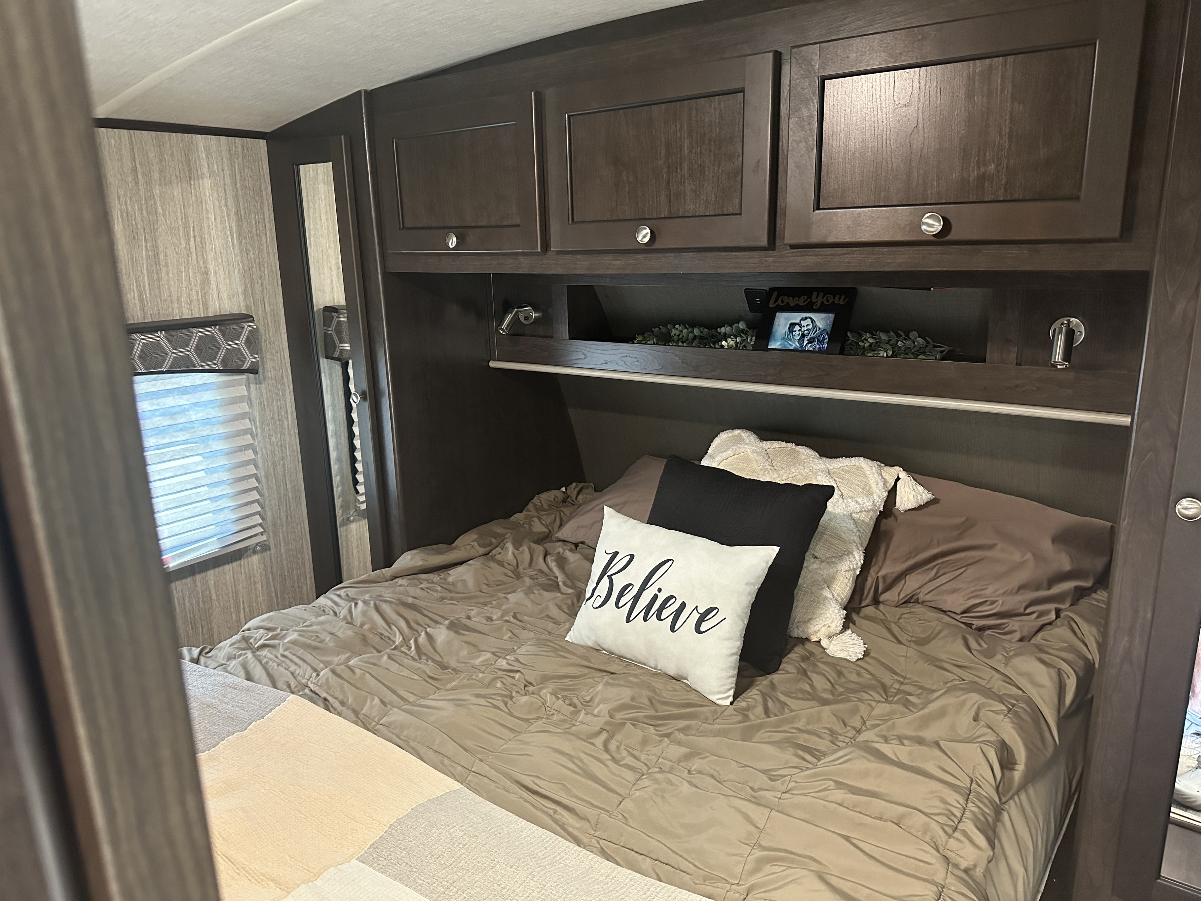 Inside a comfortable RV rental bedroom featuring dark wood cabinets, a cozy bed with decorative pillows, and a window.. Cruiser RV Shadow Cruiser 2020
