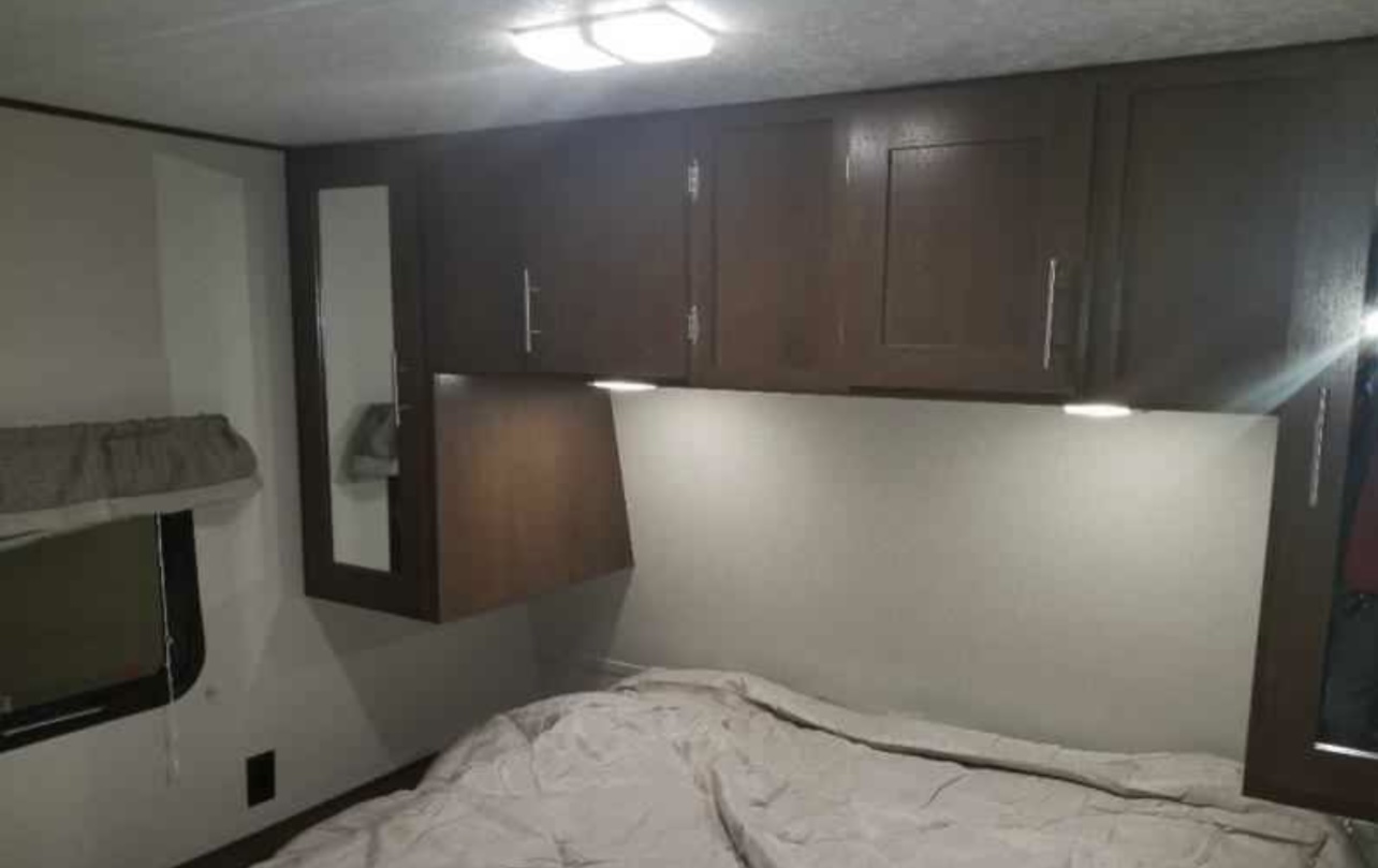 Cozy RV rental bedroom featuring dark wood overhead cabinets, bright lighting, and a comfortable bed, ideal for travel.. Avenger ATI 2018