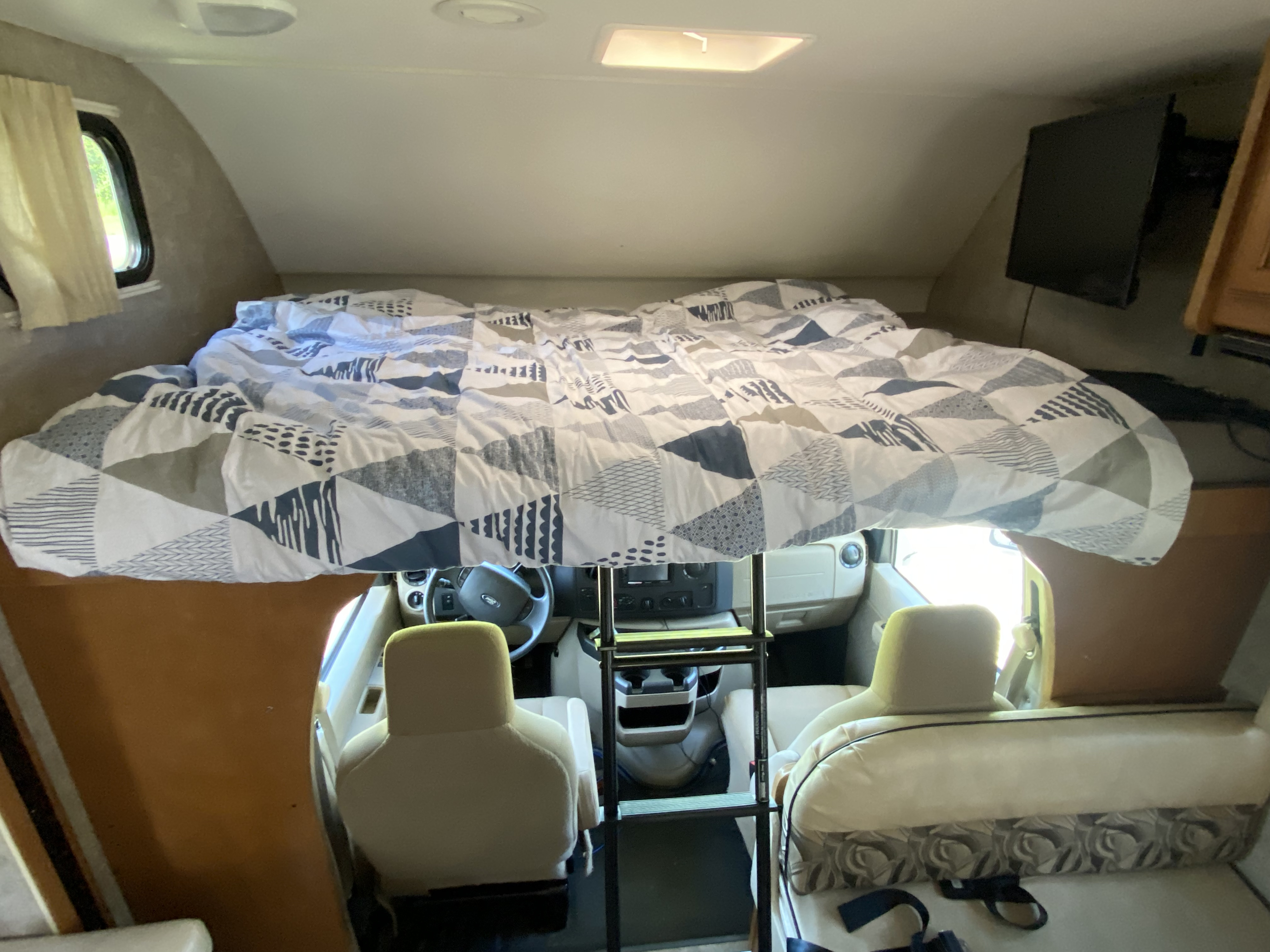 Overhead sleeping area and front seats in an RV rental. Features a bed with patterned bedding, a ladder, and a side window.. Winnebago Minnie Winnie 2015