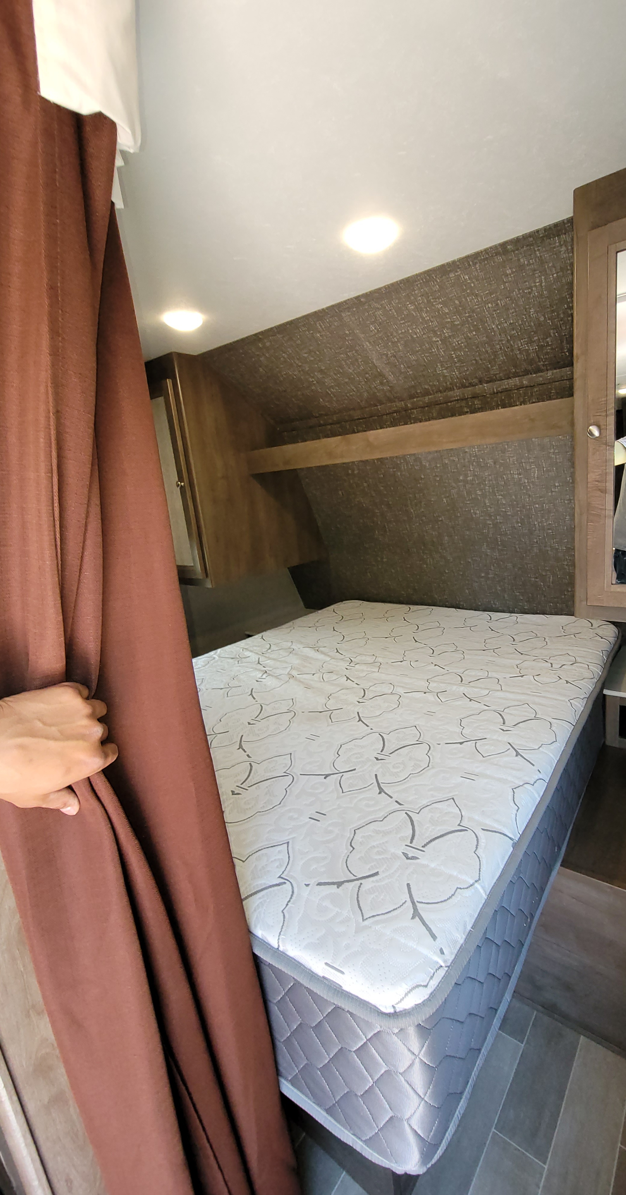 Spacious RV rental bedroom with patterned mattress, brown curtain, and overhead storage.. Dutchmen Coleman Lantern 2019