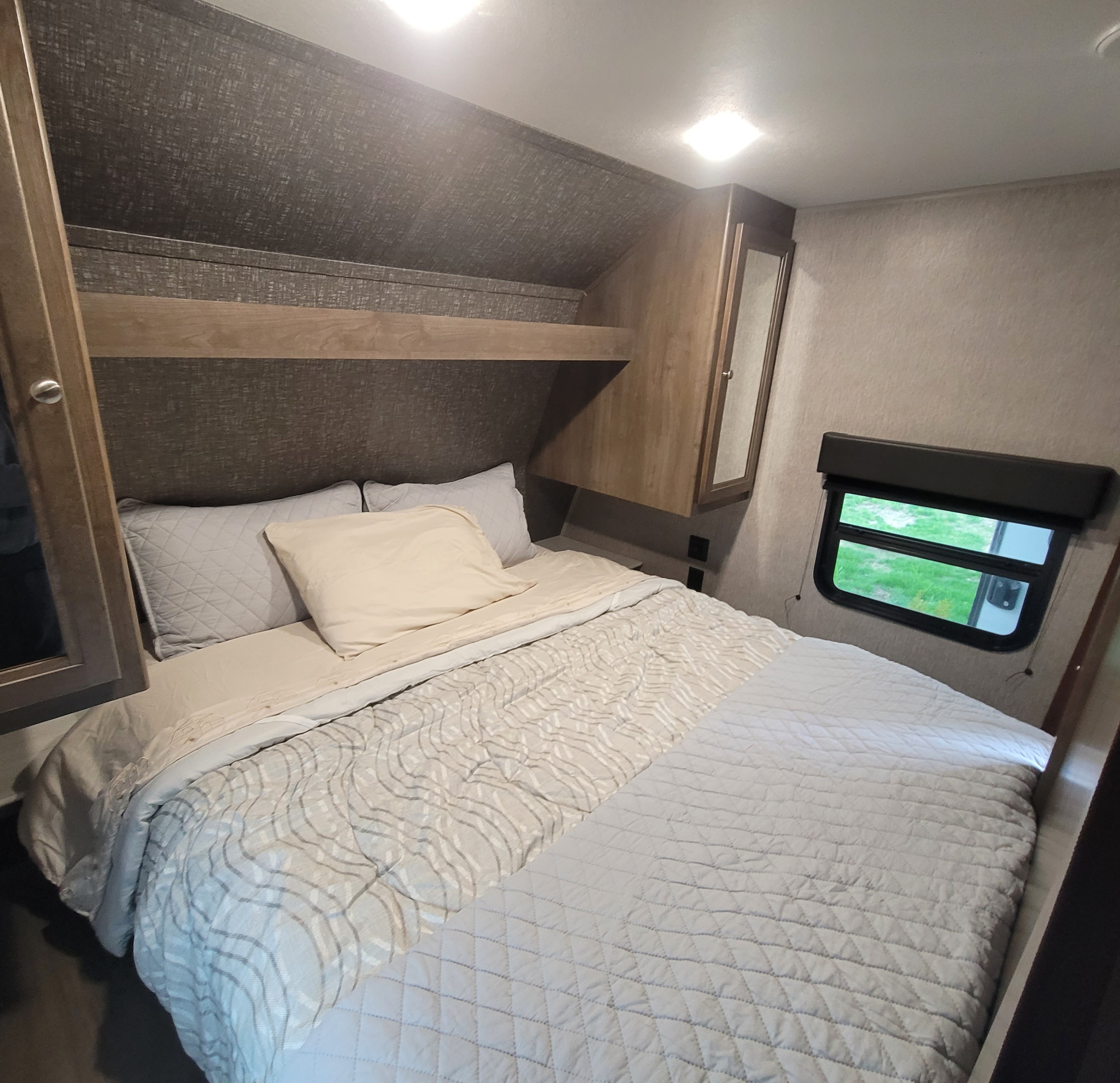 RV rental master bedroom with a light-toned bed, quilted comforter, overhead storage, and side window.. Dutchmen Coleman Lantern 2019