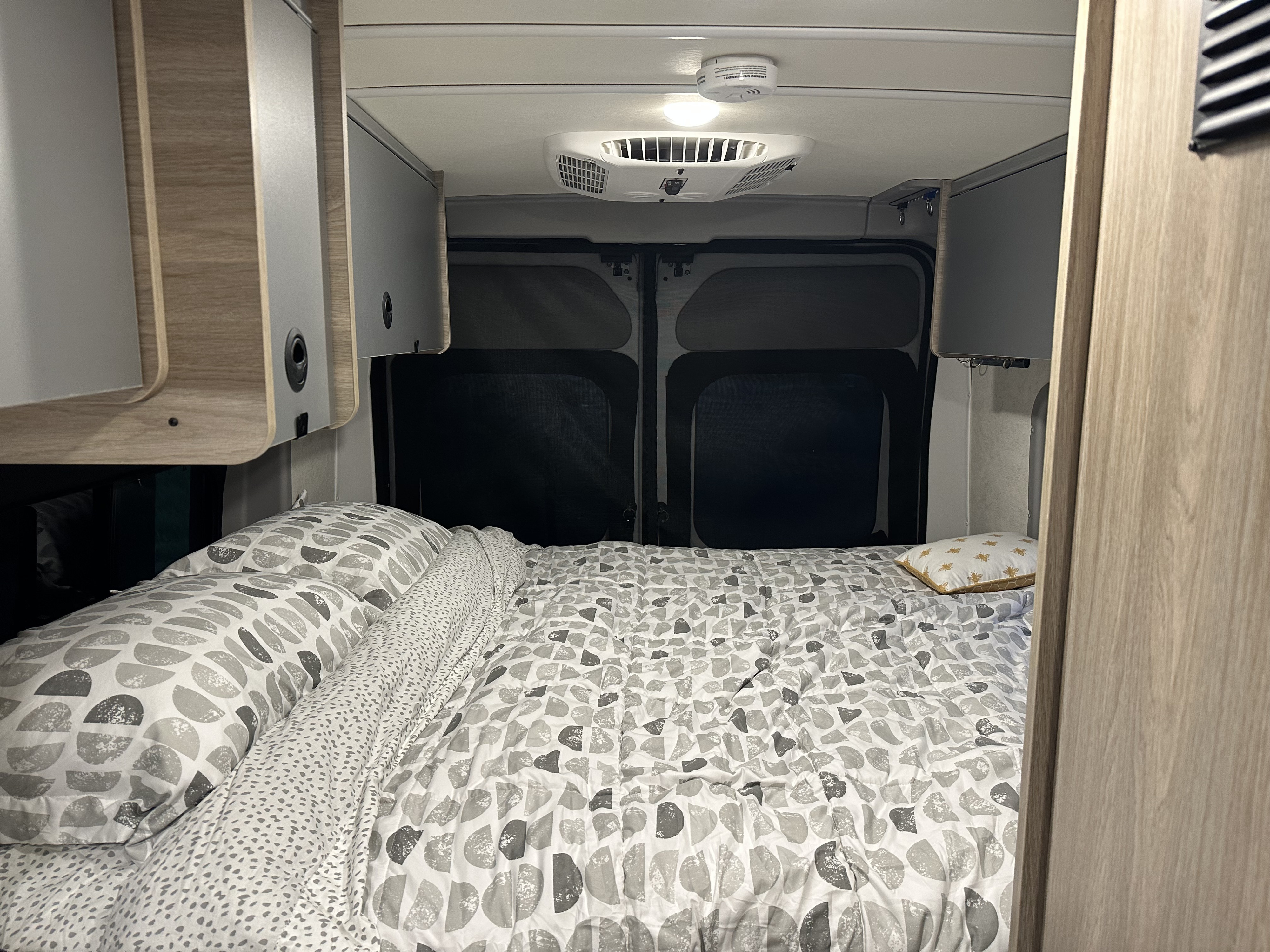 RV rental's cozy bedroom features patterned bedding, sleek overhead cabinets, and dark rear window screens for privacy.. Winnebago Solis 2023