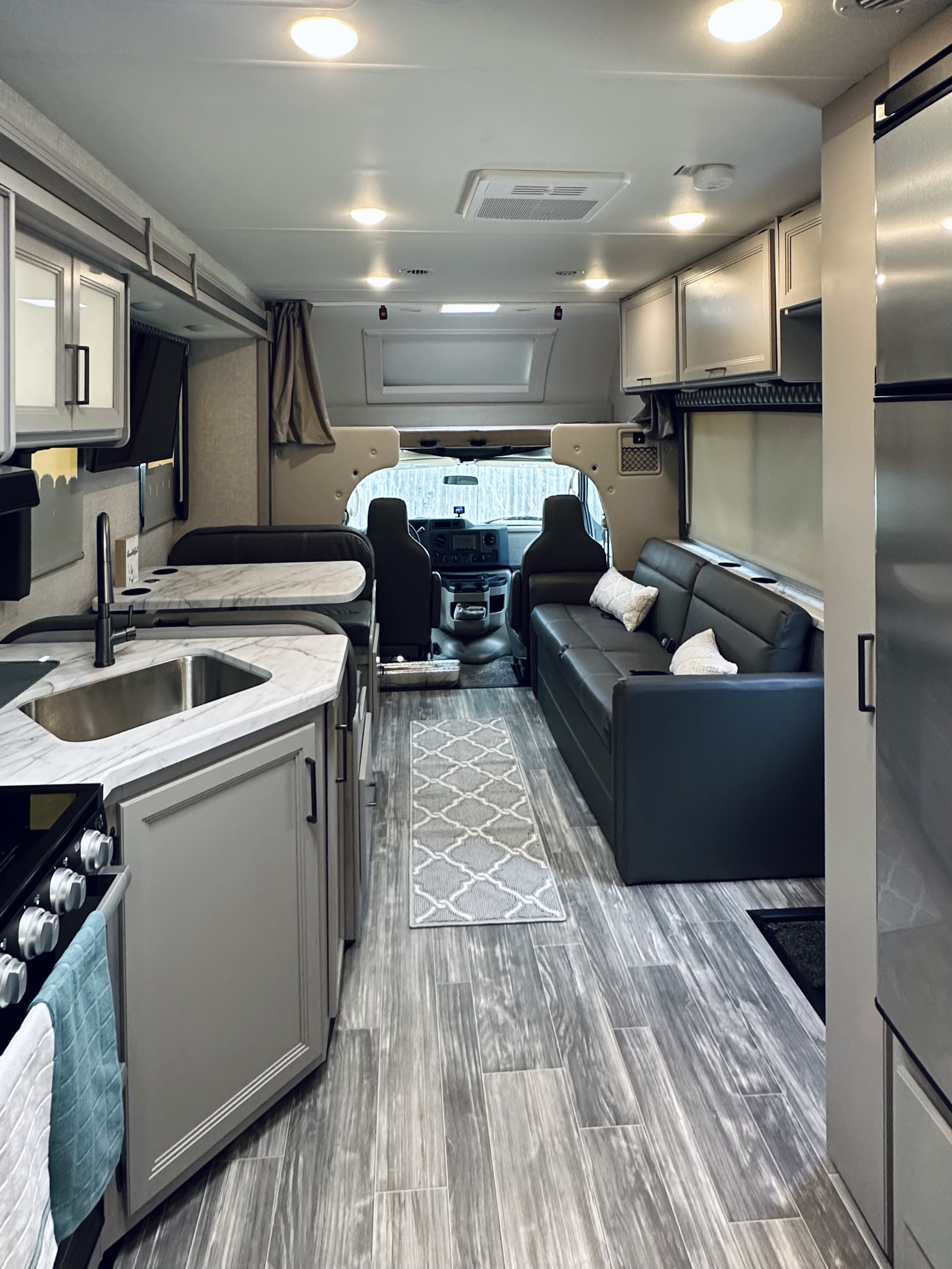 Inside a modern RV rental: a bright living space with a kitchen, dinette, sofa, and overhead bunk, all in shades of gray.. Thor Geneva 2023