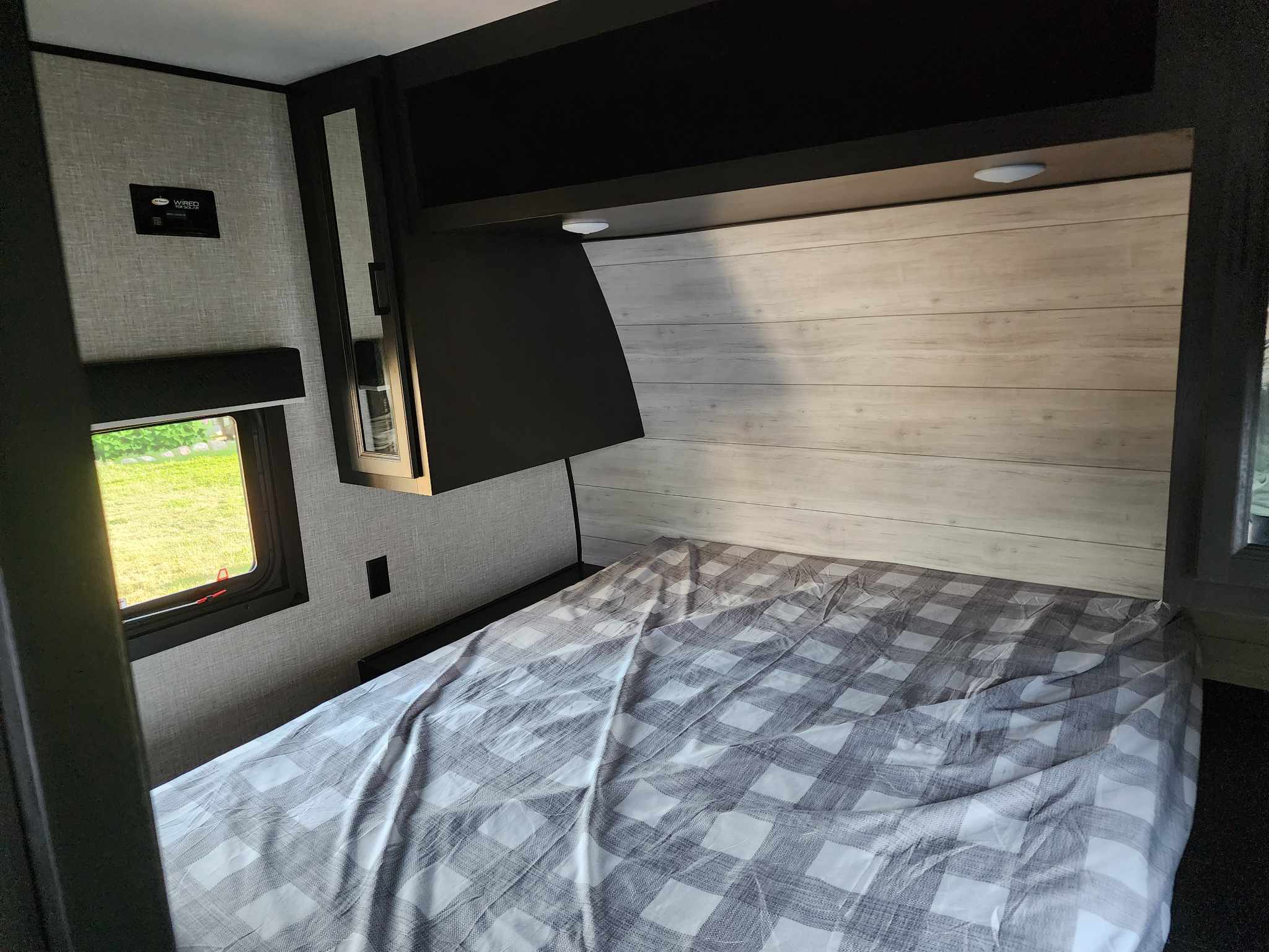 RV rental bedroom featuring a checkered bed, compact overhead storage, and a bright window view.. Jayco Jay Flight 2023