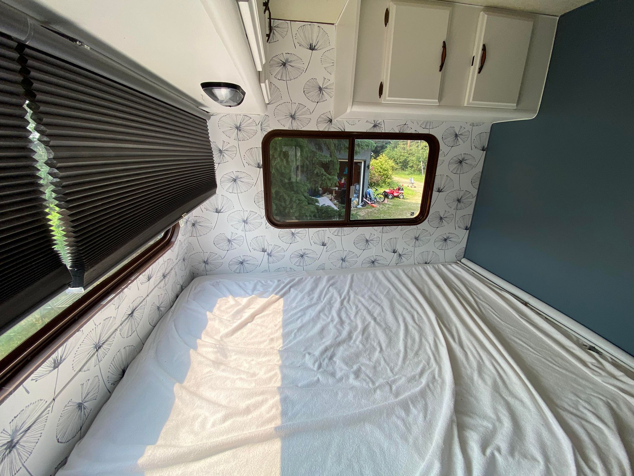 Inside an RV rental bedroom: white bed, dandelion wallpaper, window view with greenery, dark blinds & blue accent wall.. Frontier Ford Econoline 1986