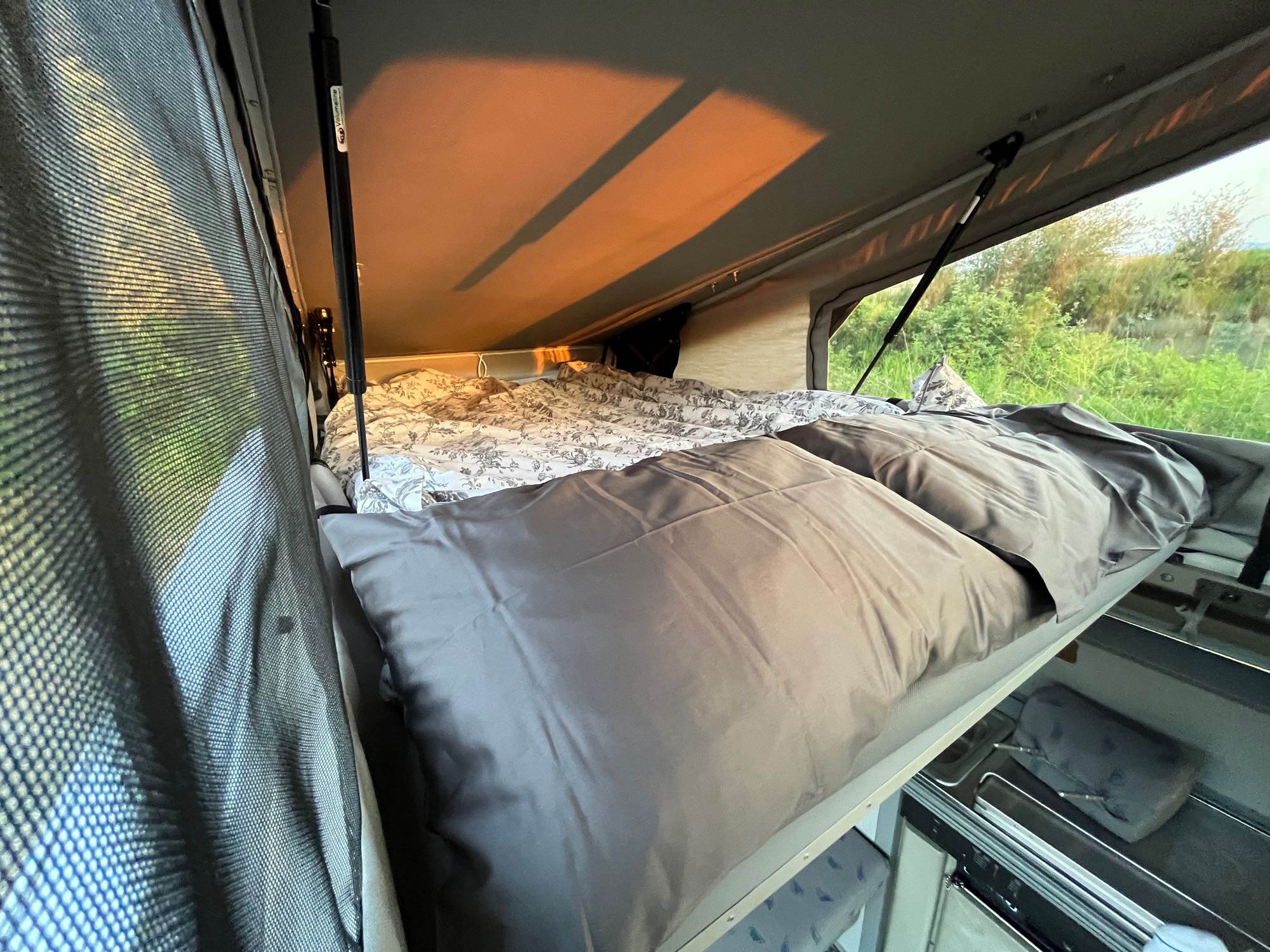 Cozy RV rental pop-top bed with patterned sheets and warm lighting, offering peaceful nature views.. Volkswagen Eurovan Winnebago 1997