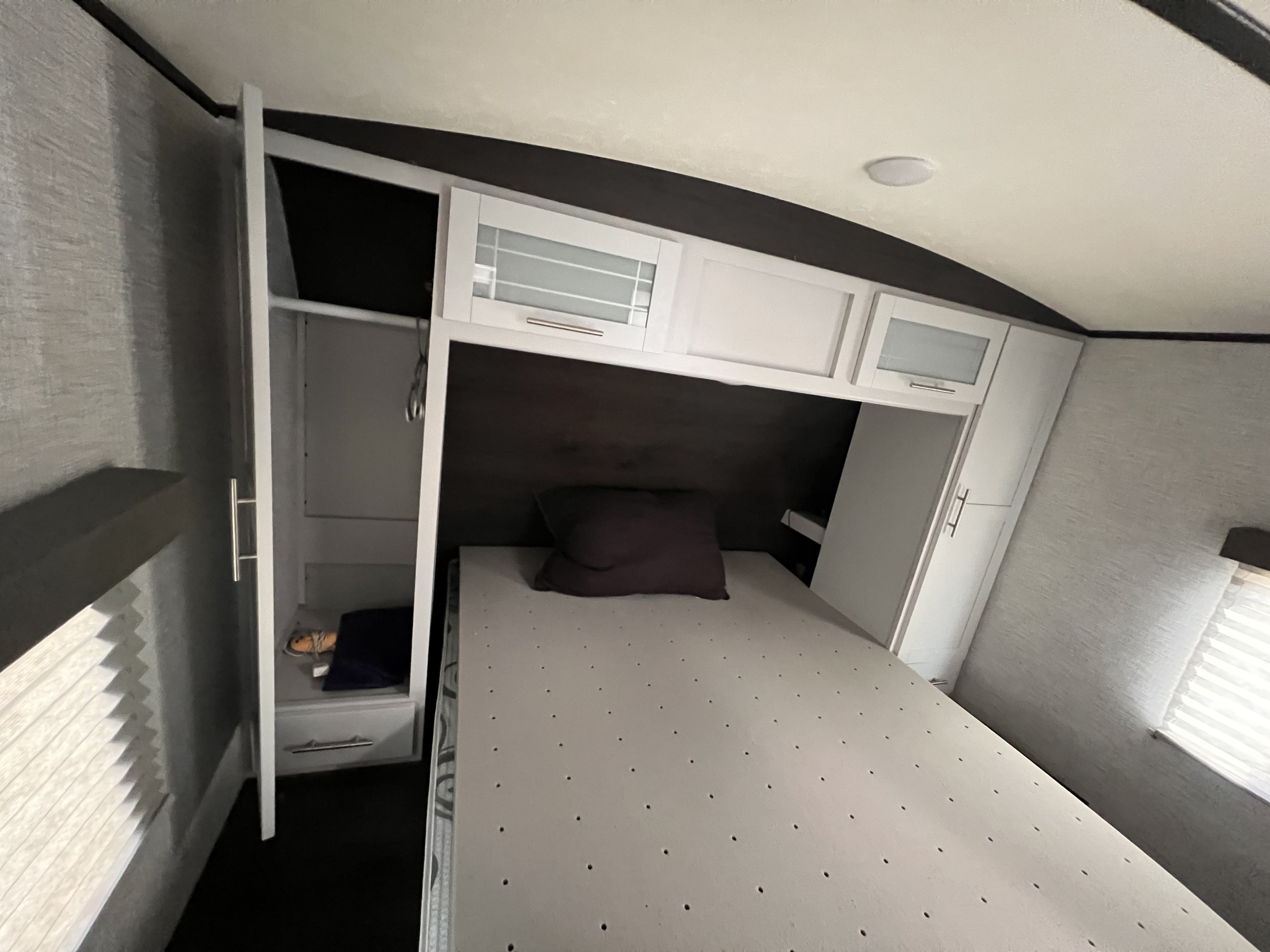 Interior of an RV rental bedroom featuring a bed, brown pillow, white overhead cabinets, and an open wardrobe. Textured grey walls.. Keystone Bullet 2021