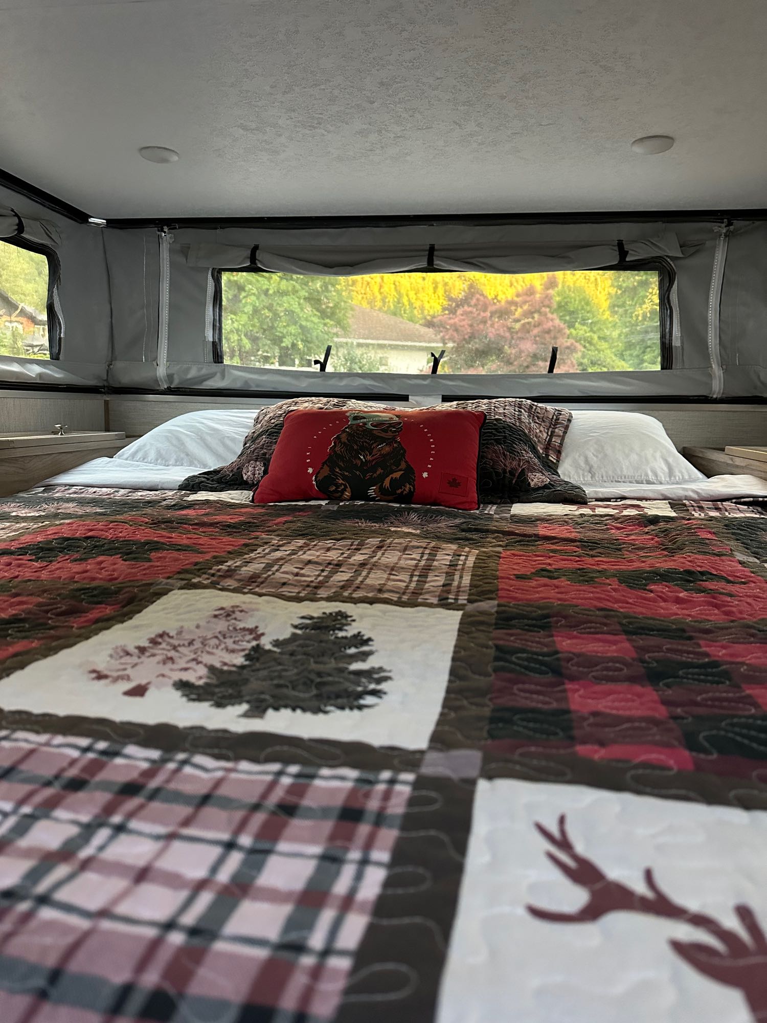 Cozy RV rental bed with a rustic quilt, bear pillow, and scenic outdoor view through the windows.. Forest River Travel lite 2022