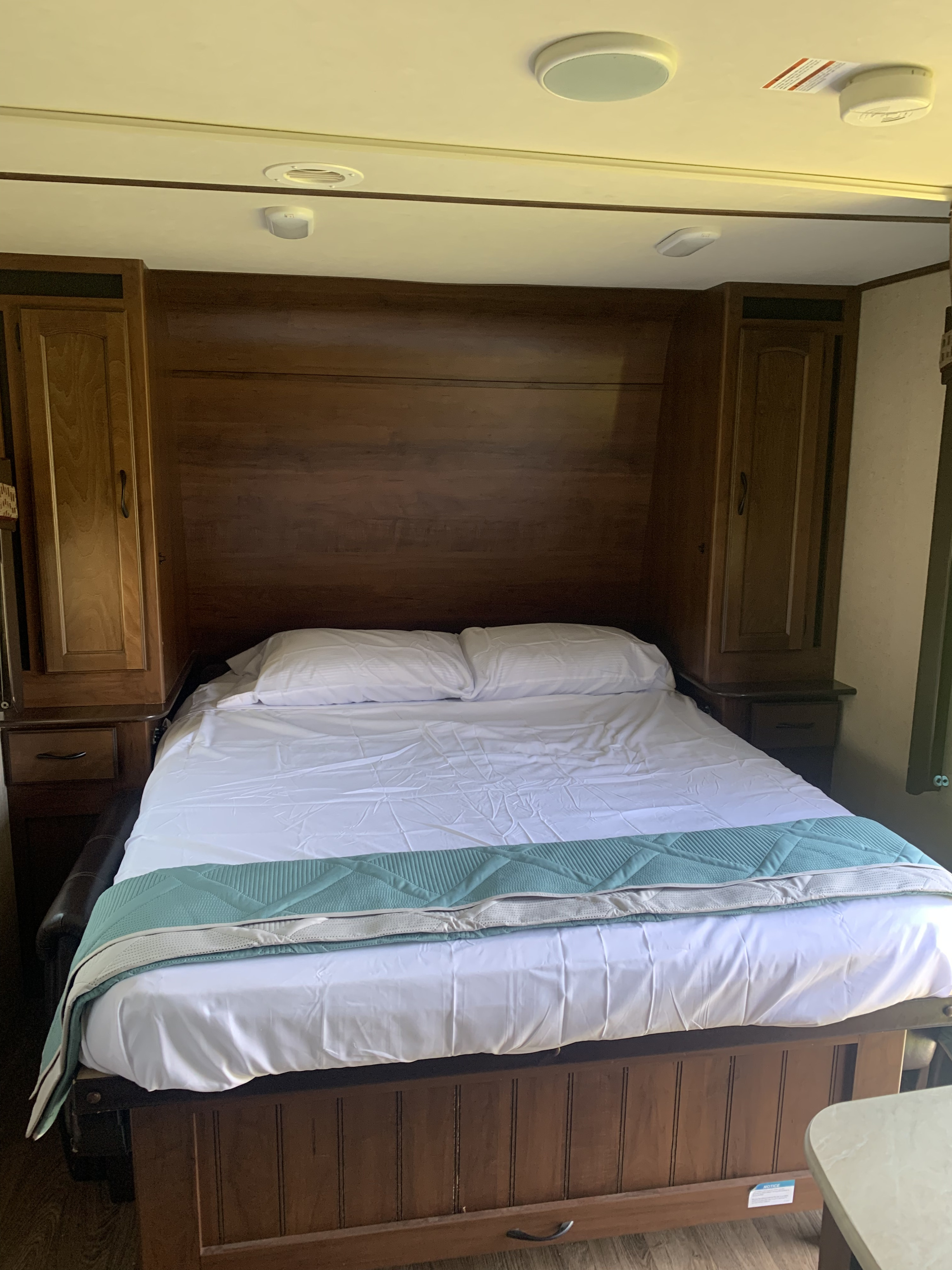 Inside an RV rental: a neatly made queen bed with white linens and a teal accent blanket, flanked by wood cabinetry.. Jayco White Hawk Ultra Lite 2015
