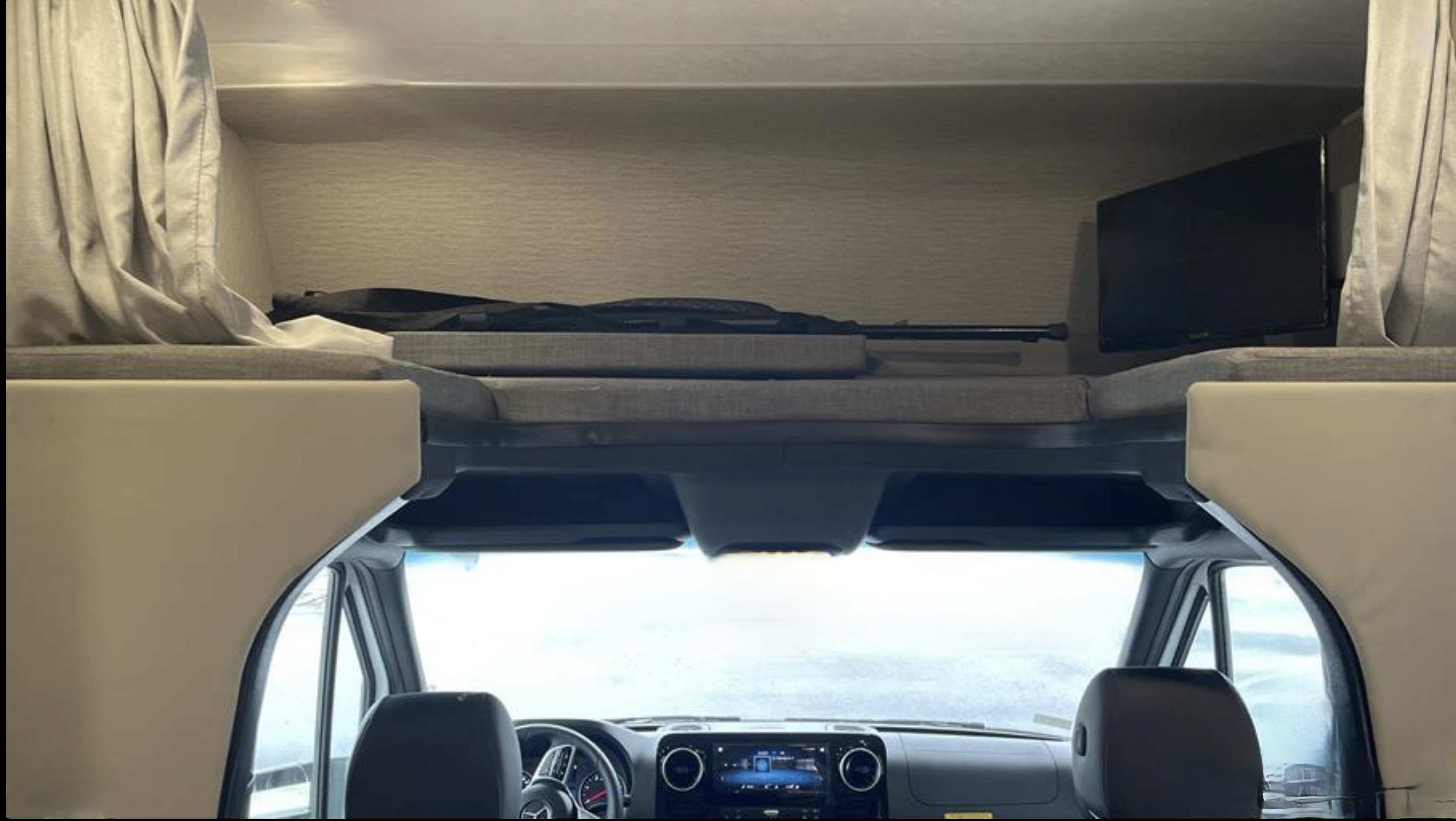 Interior of an RV rental: A cab-over bunk bed with a TV, curtains, and a rifle, looking down at the driver's dashboard.. Coachmen Prism 2023