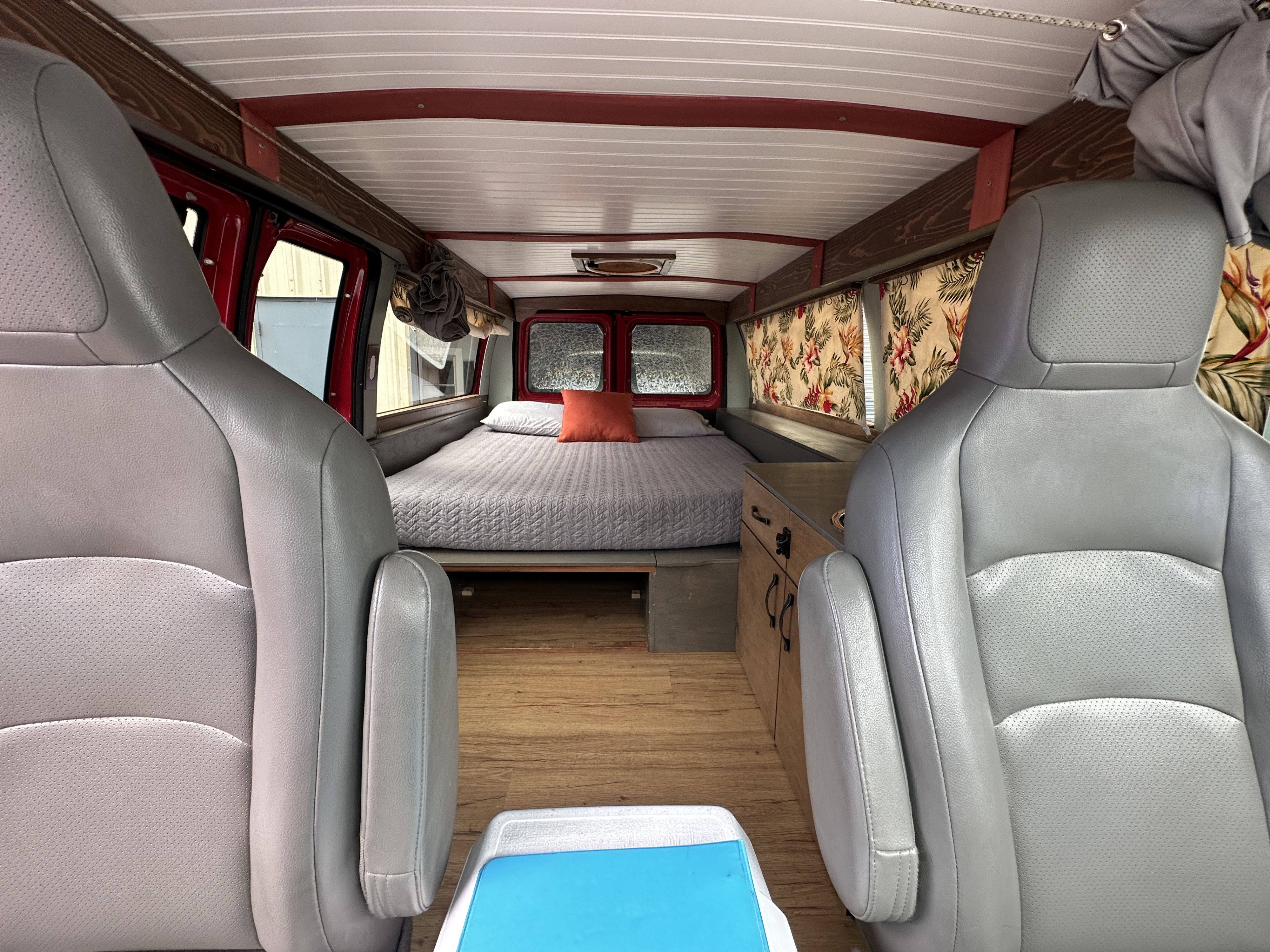 Cozy RV rental interior: grey seats face a comfy bed, wood floors, and a small kitchenette with floral curtains.. Ford E150 2013