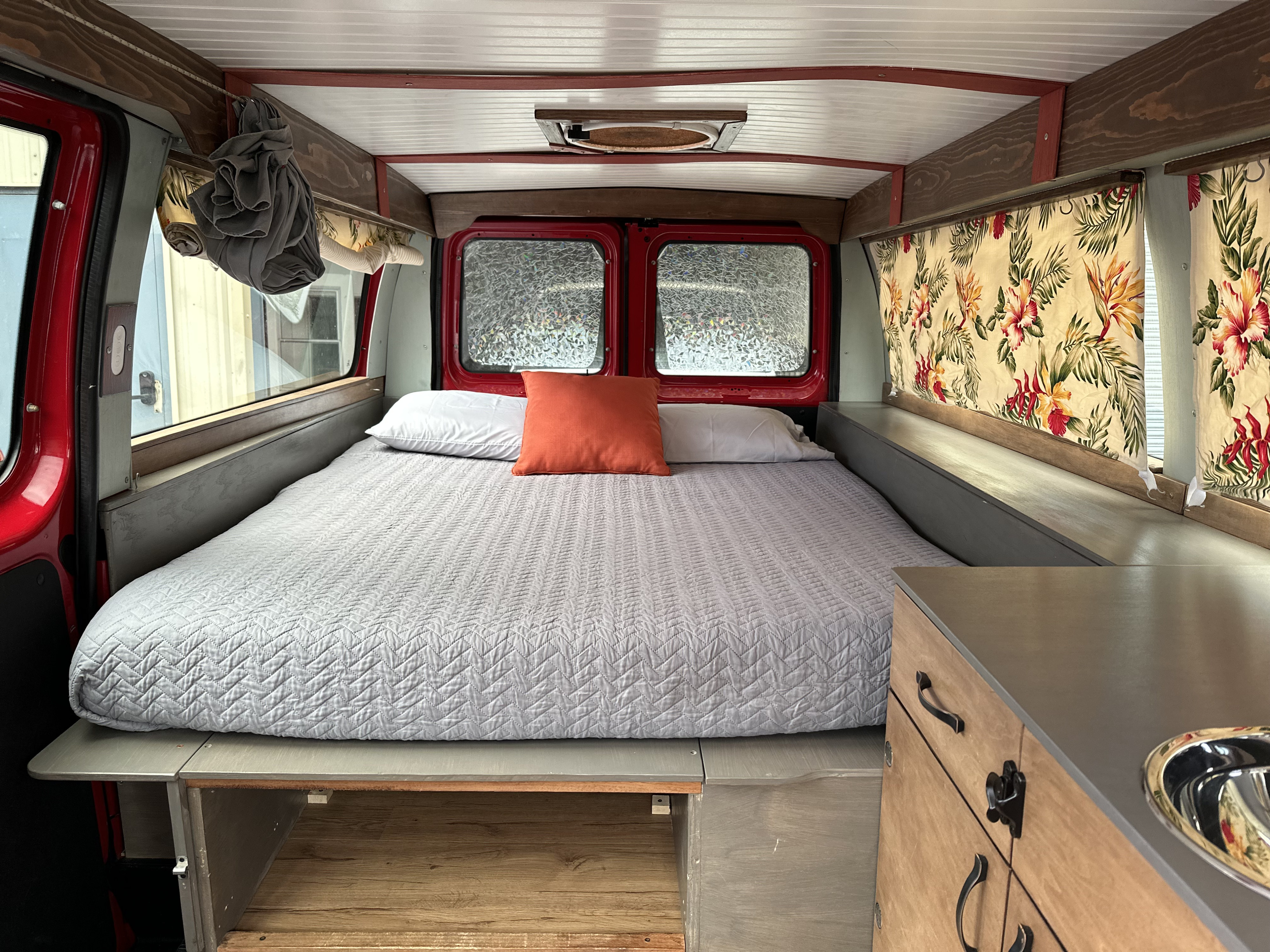 An RV rental's clever interior design shows a comfortable bed, floral curtains, and a compact kitchen counter with sink.. Ford E150 2013