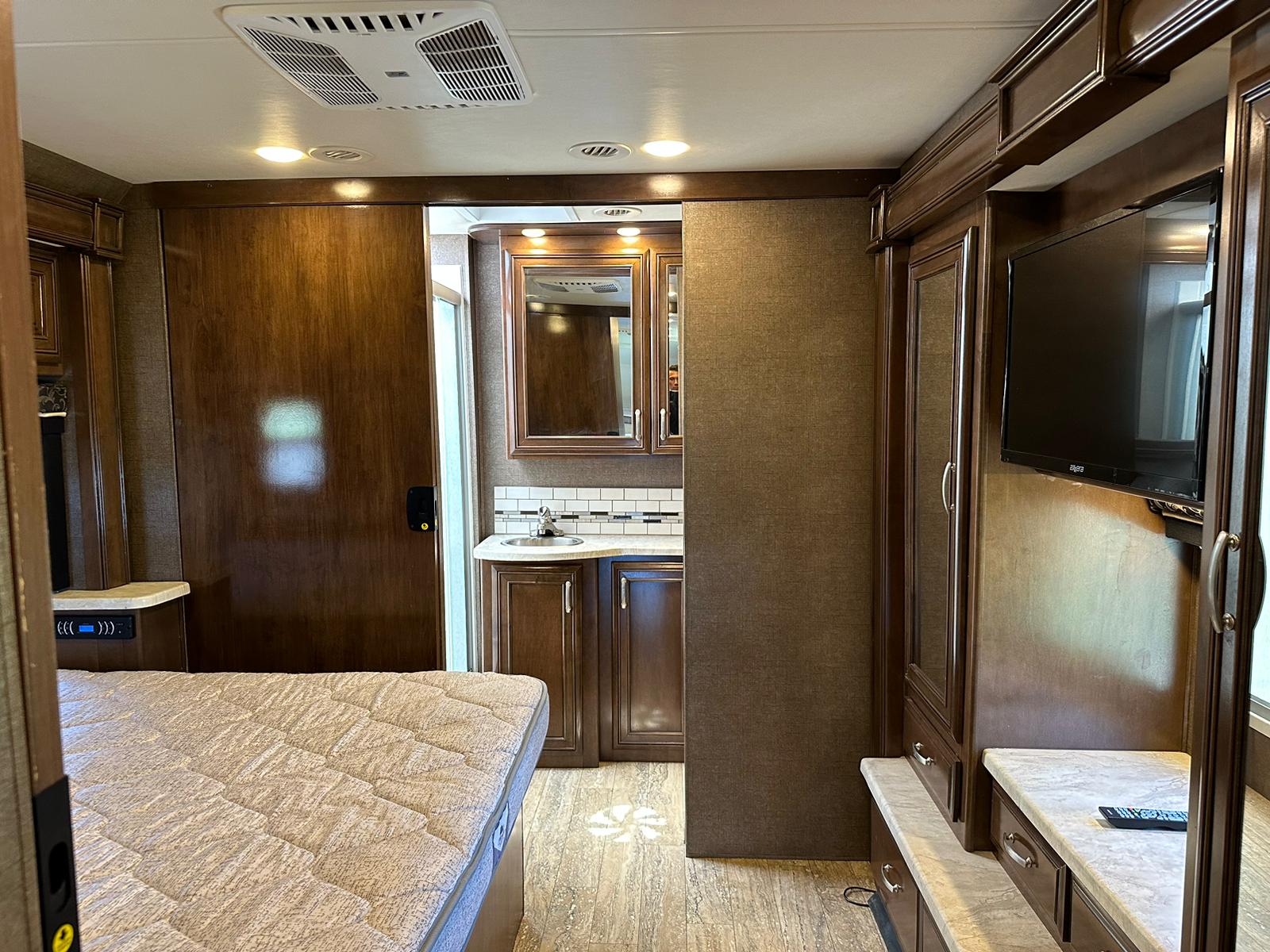 Spacious RV rental interior: bedroom with bed, TV, and connected bathroom vanity. Dark wood cabinetry & light flooring.. Thor Four Winds 2017