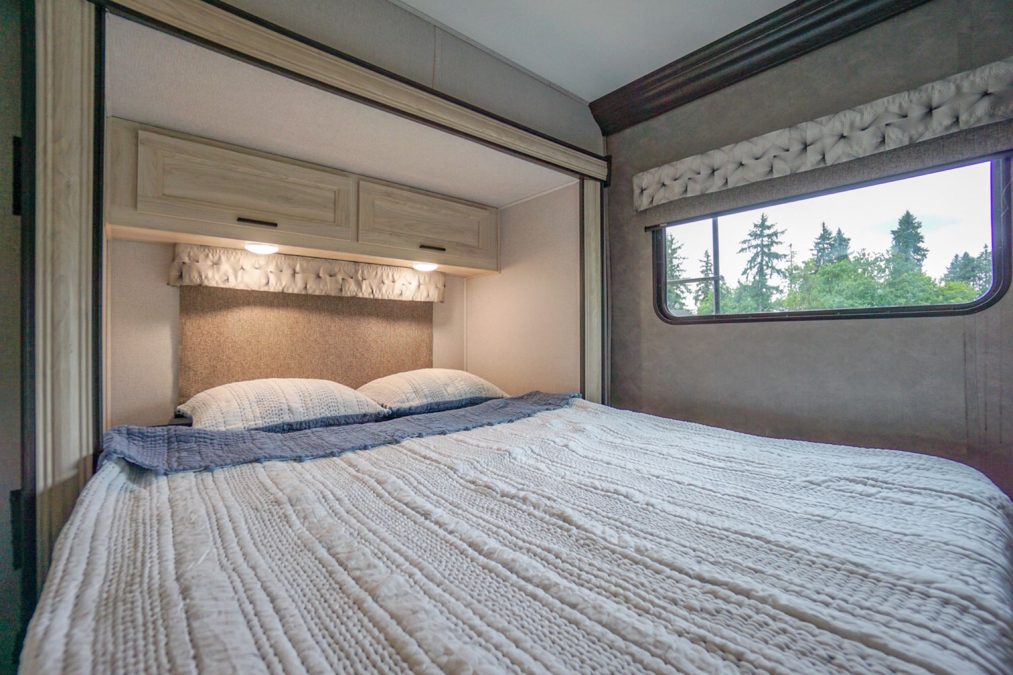Cozy RV rental bedroom with a comfy bed, overhead cabinets, and a window showing green trees outside.. Forest River Sunseeker LE 2020