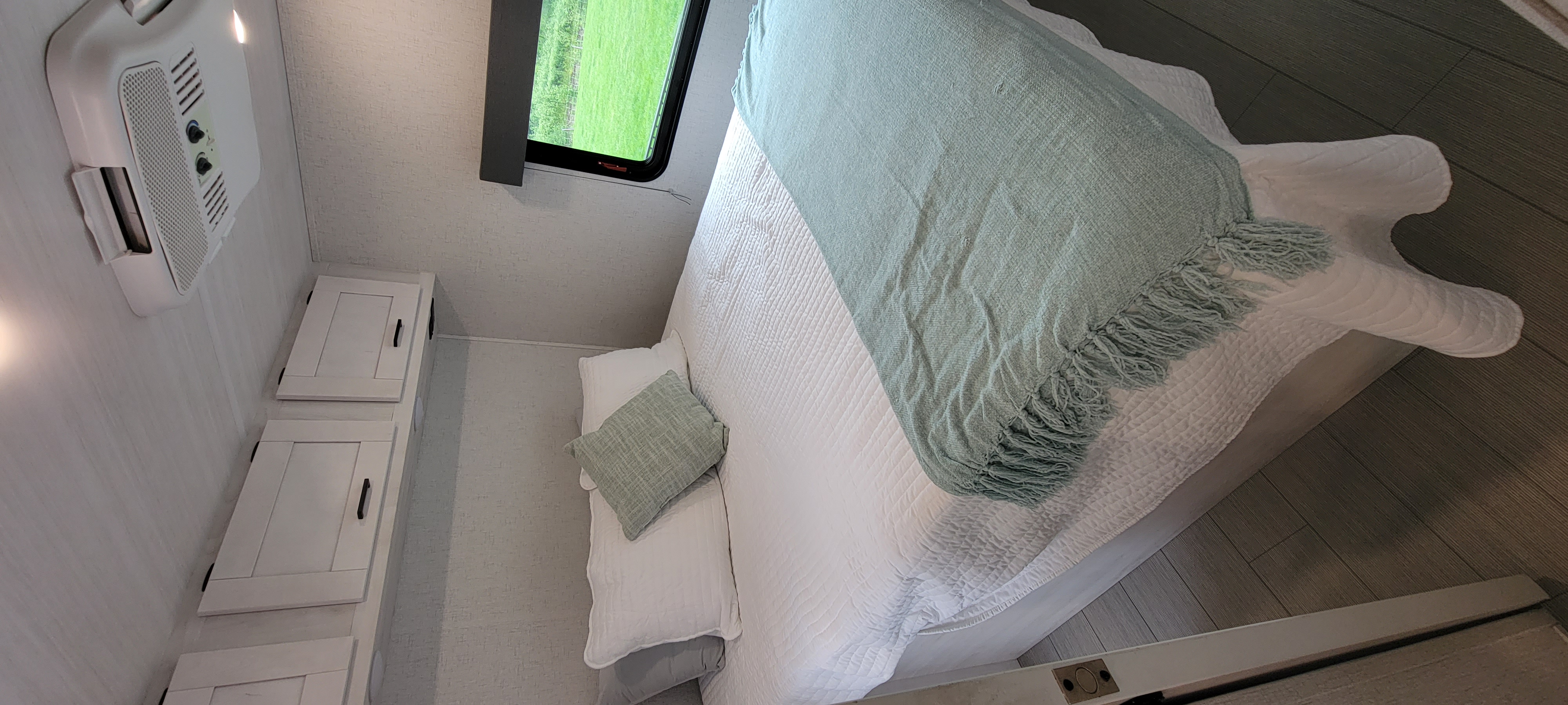 Cozy RV rental bedroom with white bedding, a mint throw, overhead cabinets, and AC. Features a window with a green view.. East to West Entrada 2023