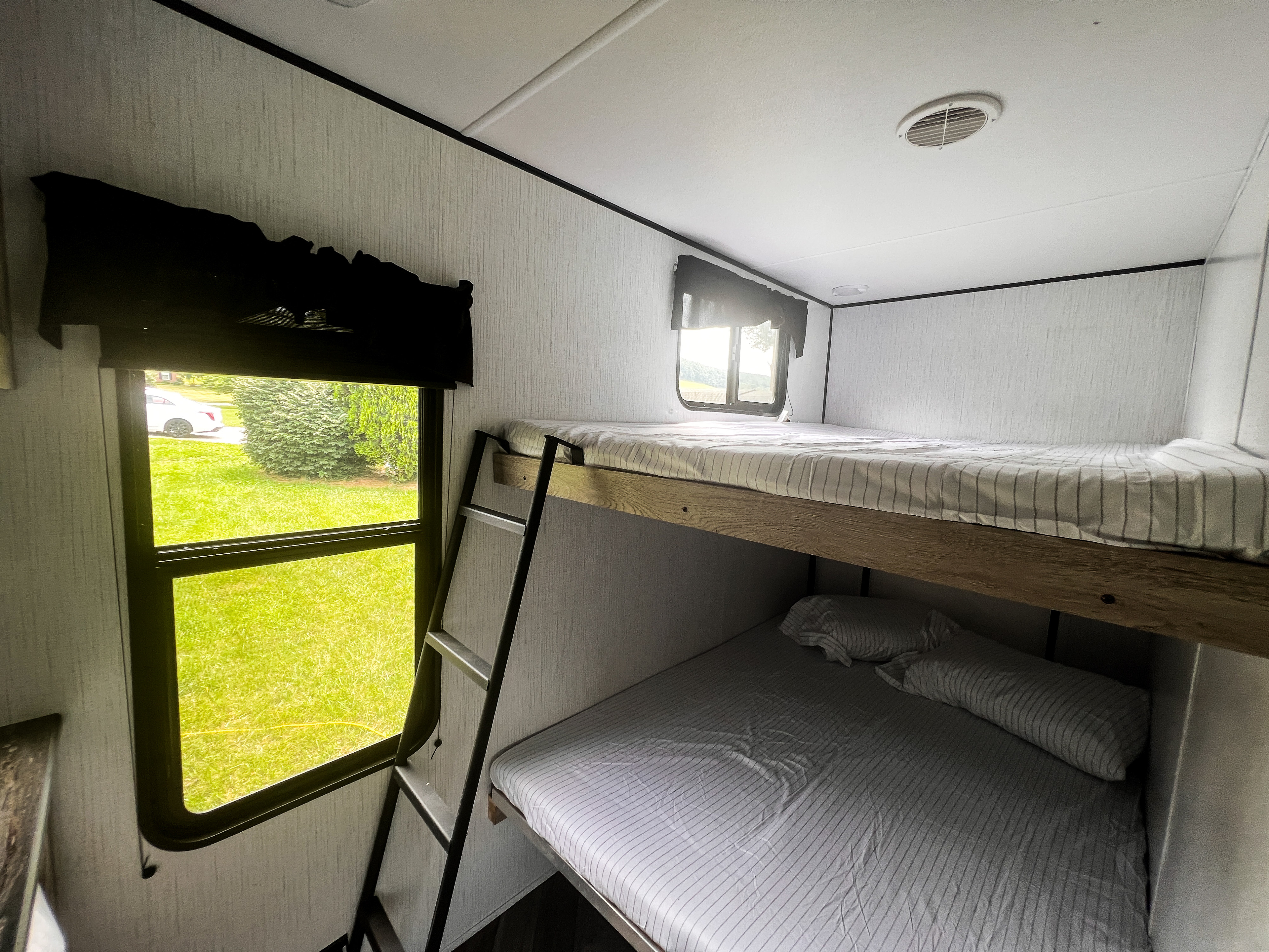 Inside an RV rental: comfortable bunk beds with striped sheets & a ladder. Window view of green outdoor space & car.. Heartland RVs Prowler 2022