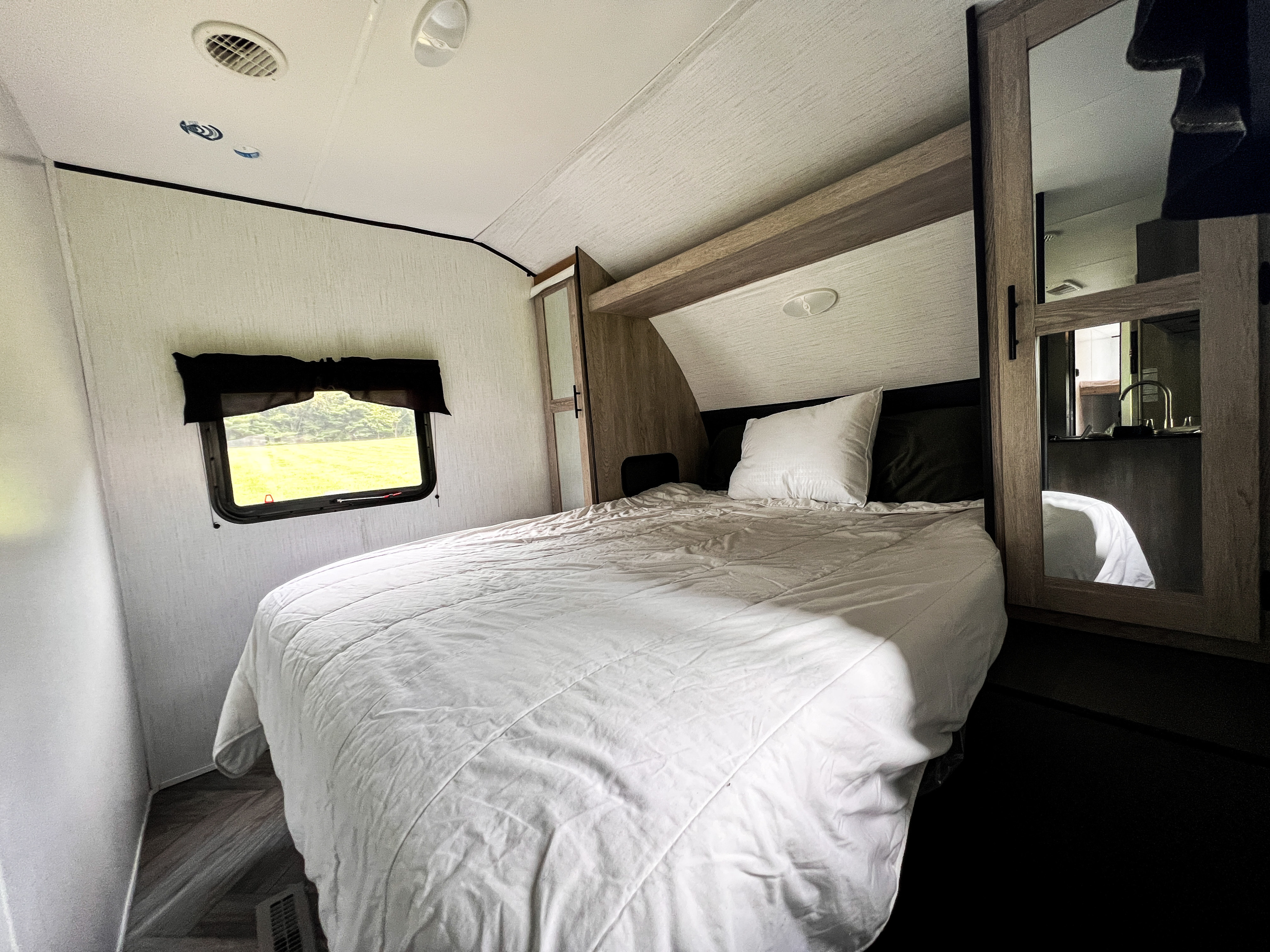 Cozy RV rental bedroom with bed, window showing greenery, and a mirrored wardrobe reflecting the interior kitchenette.. Heartland RVs Prowler 2022