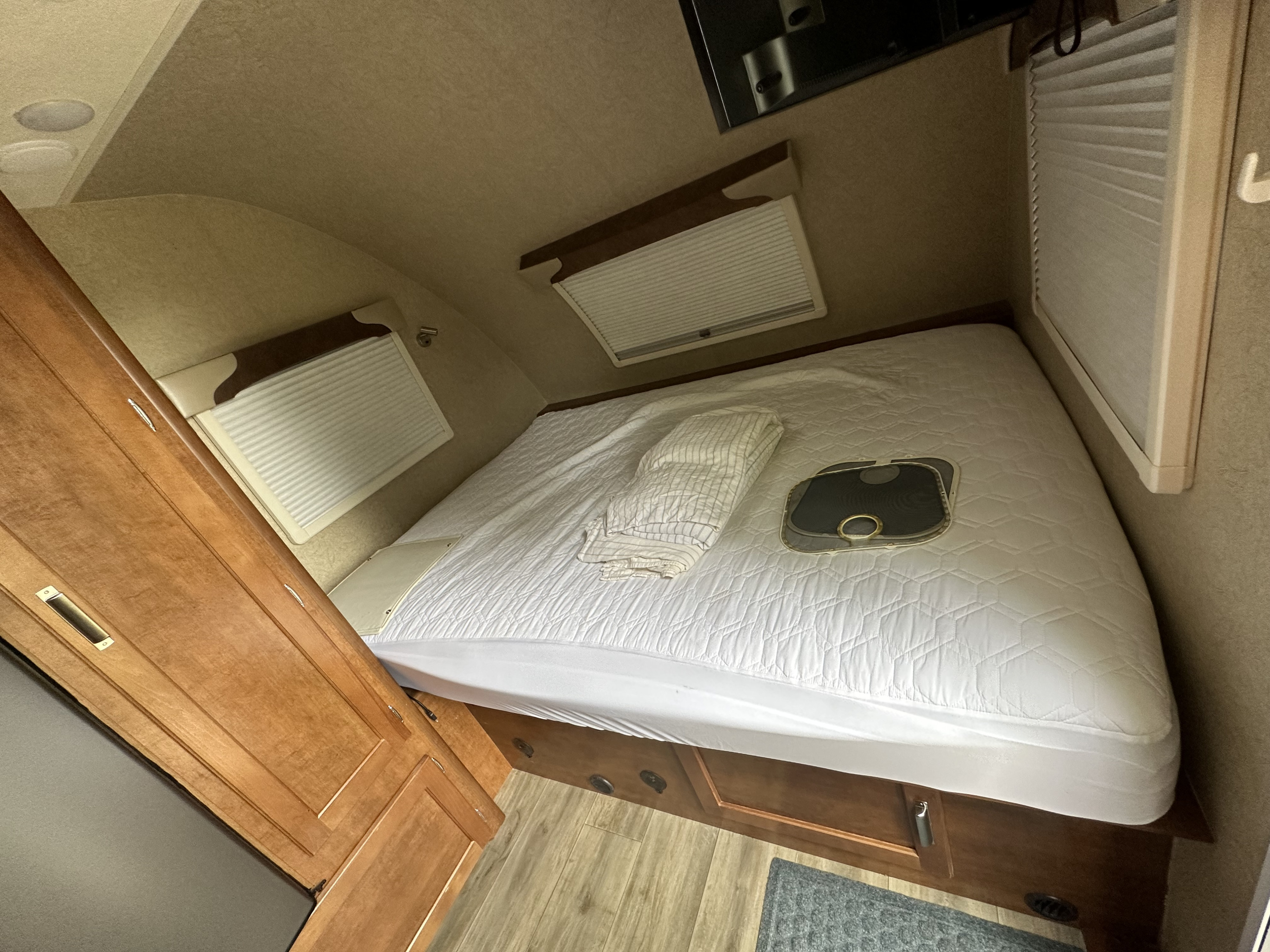 Cozy RV rental interior featuring a bed with a white quilted cover, wood cabinetry, and pleated window blinds.. Lance Manufacturing Lance Manufacturing Trailer 2020