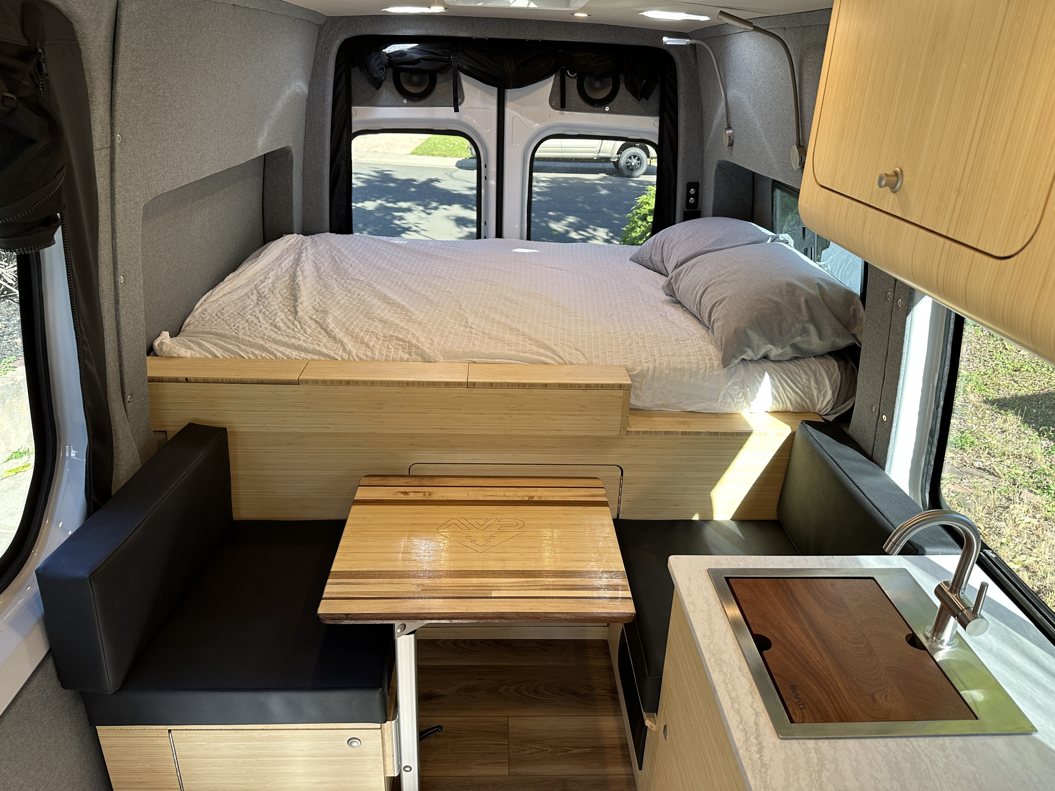 Interior of a modern RV rental camper van with a bed, bamboo table, and compact kitchen featuring a sink.. Mercedes-Benz Sprinter 2019