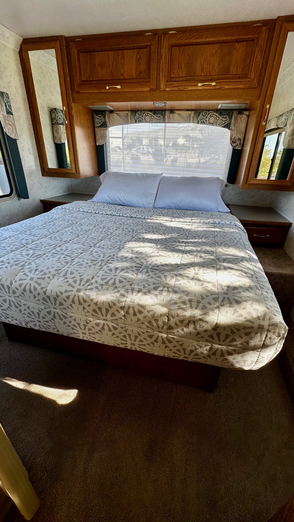 Cozy RV rental bedroom featuring a comfortable bed with a patterned comforter, overhead wood cabinets, and side tables.. Four Winds Hurricane 2002