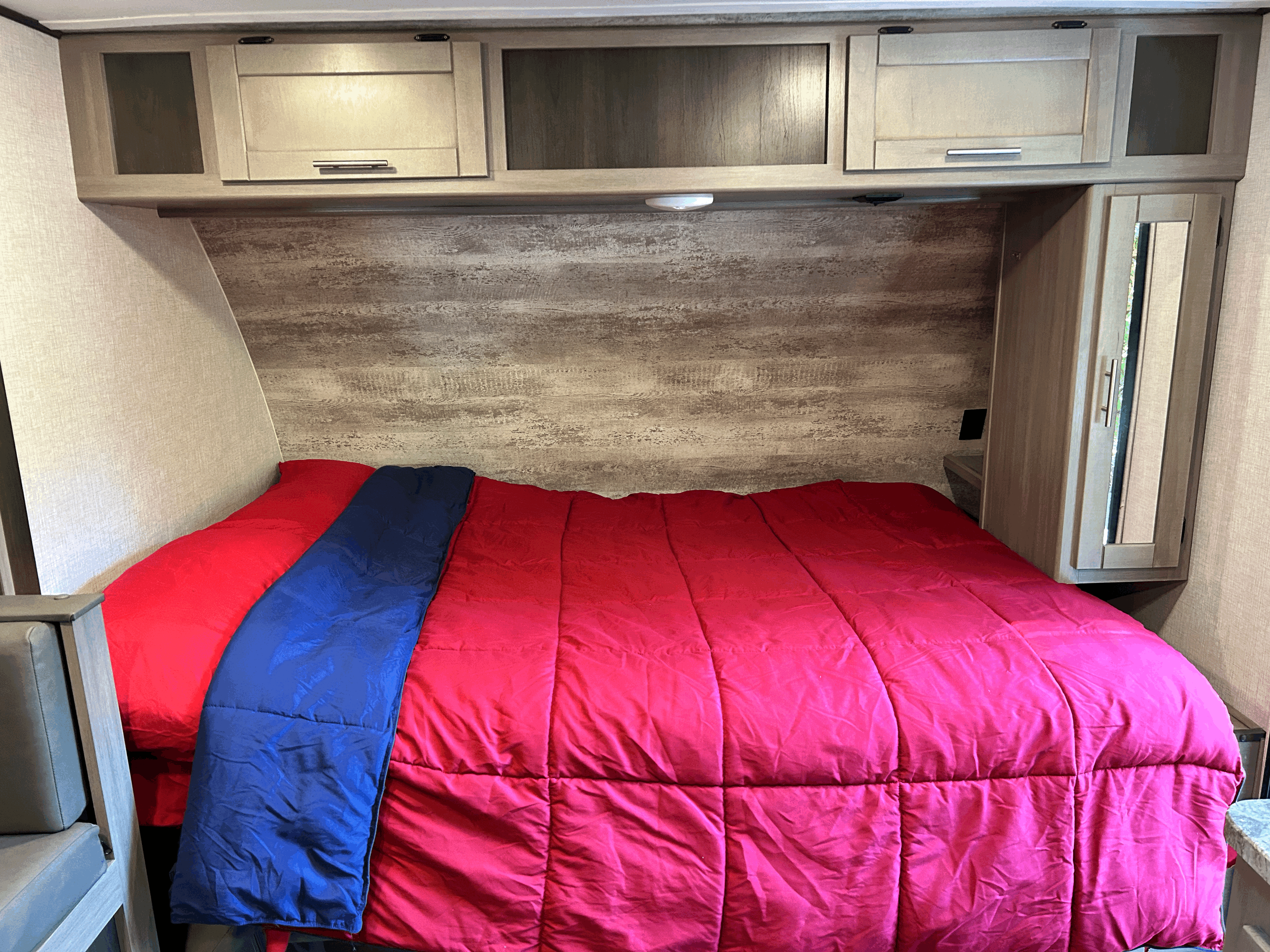 Cozy RV rental bedroom with bright red comforter, blue blanket, and light wood cabinetry.. Sunset Trail 188BH 2022