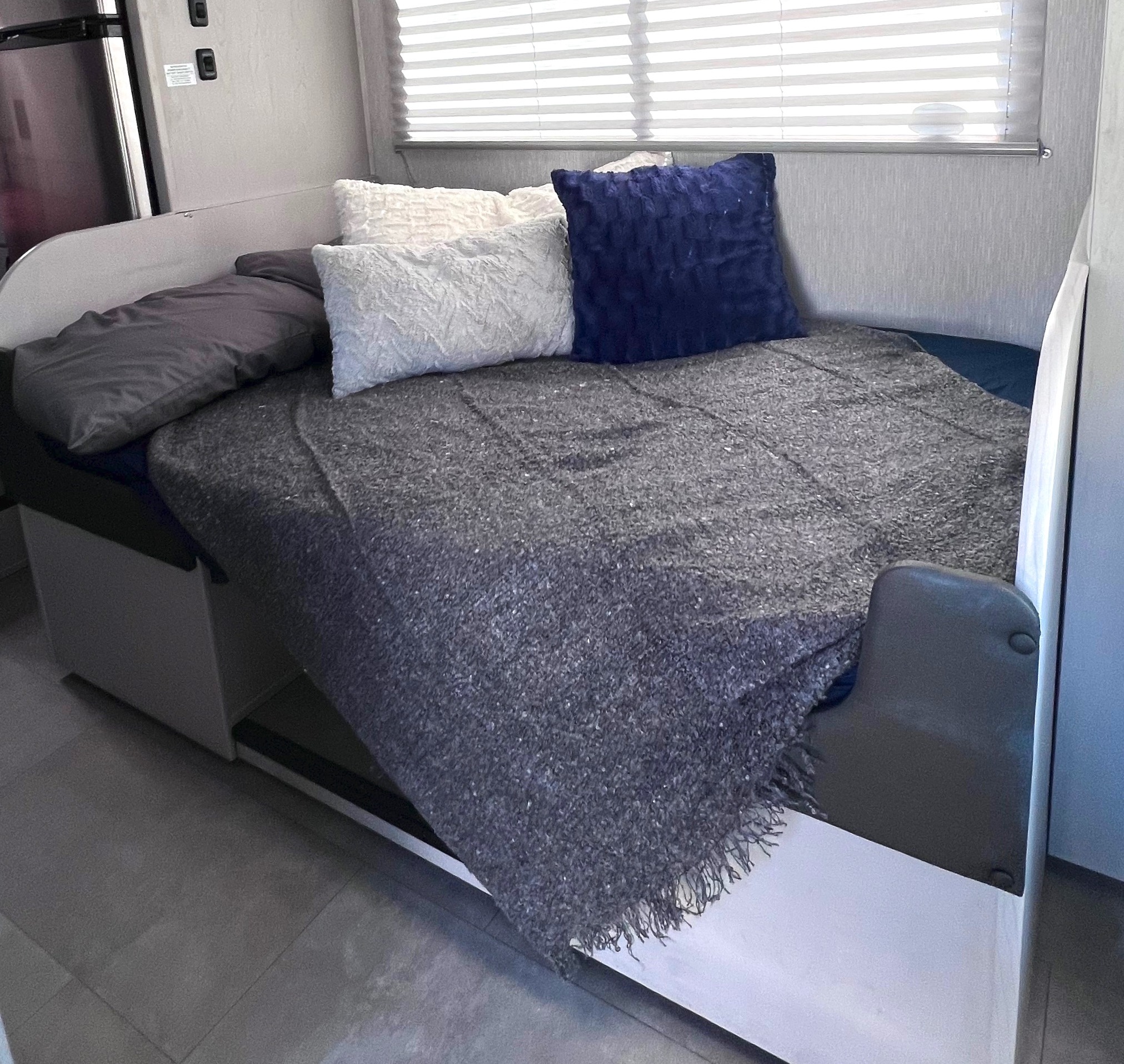Comfortable RV rental sleeping area featuring a gray blanket, navy and white pillows, and a compact fridge.. Forest River Coachmen Freelander 2023
