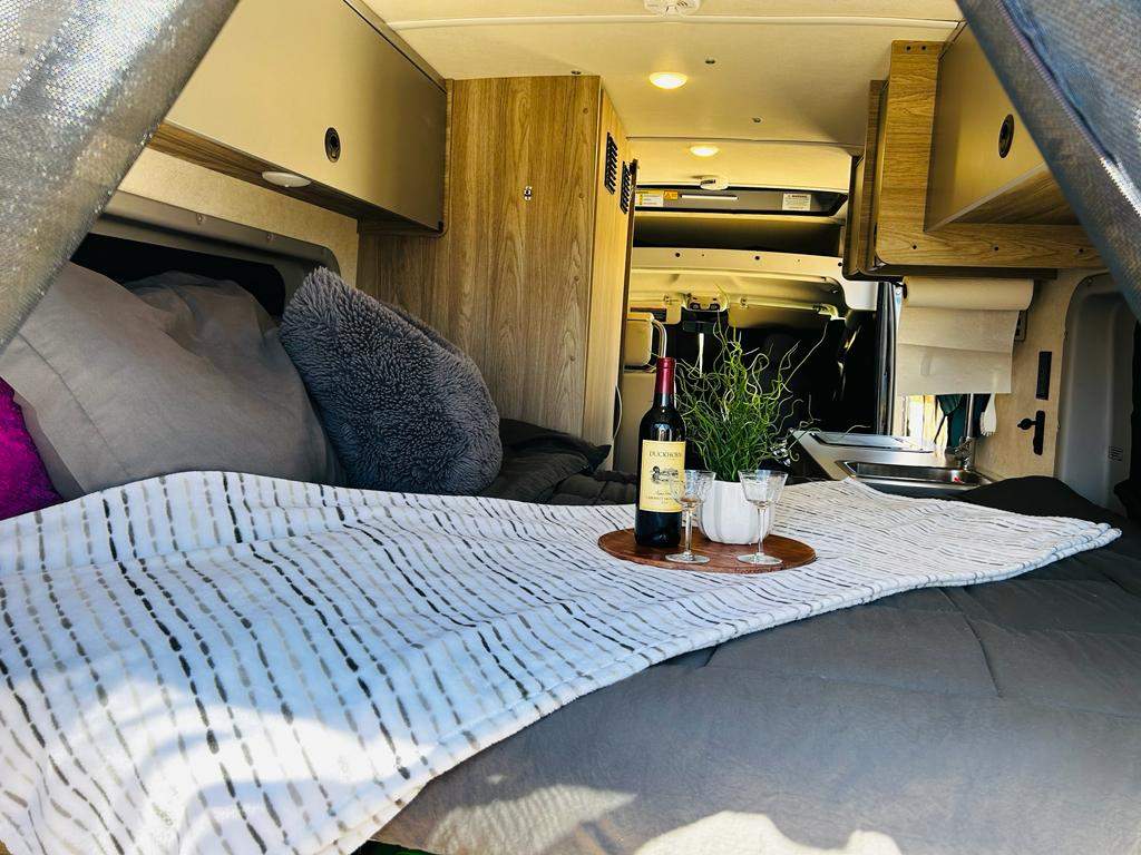 Cozy RV rental interior: bed with pillows, blanket, and a tray holding wine, glasses, and a plant for comfort.. Winnebago Solis 2023
