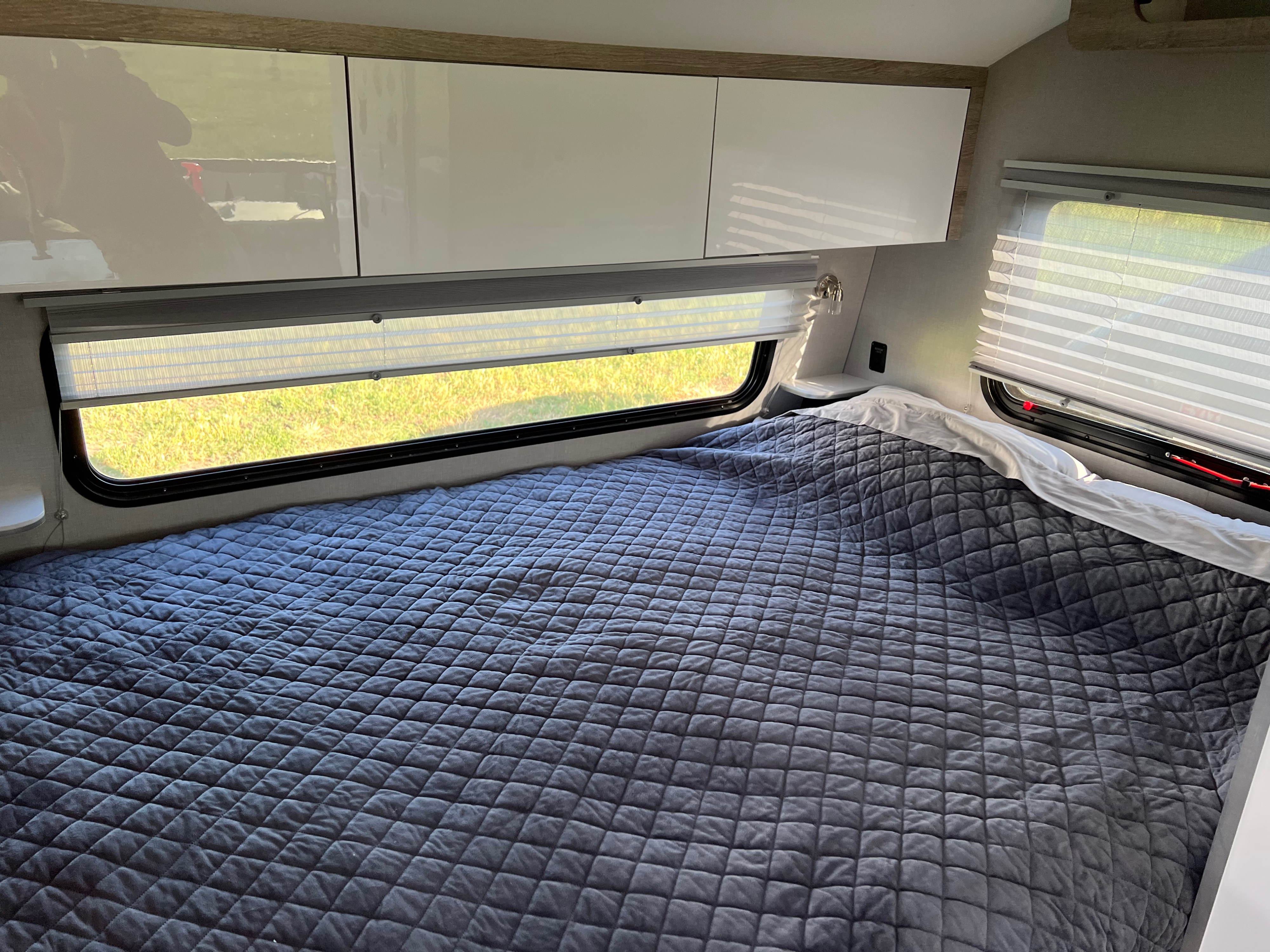 Cozy RV rental bedroom featuring a grey quilted bed, large windows with blinds, and modern overhead cabinets.. InTech RV Sol 2022