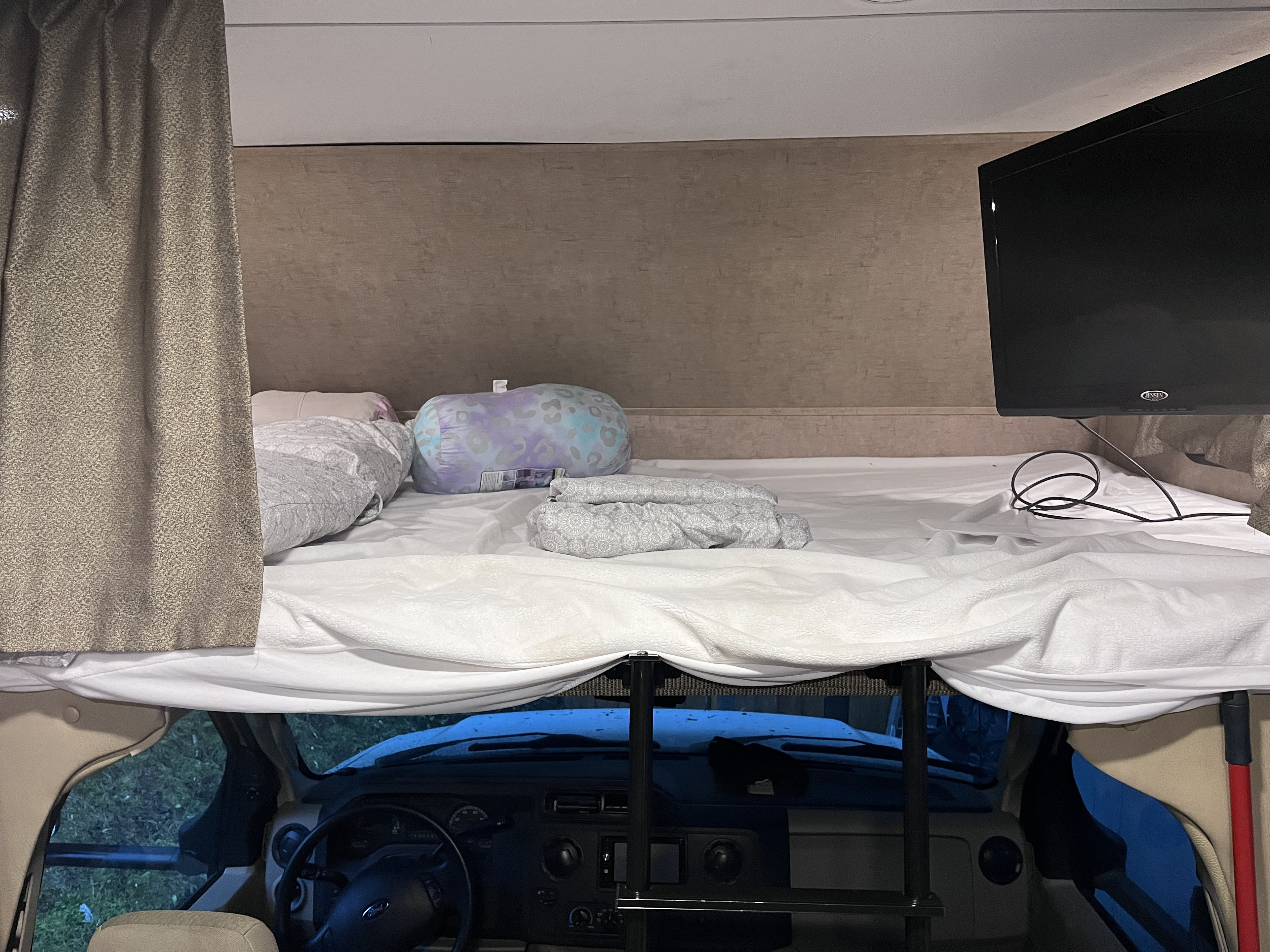 Inside an RV rental: a cozy overhead bunk bed, ready with bedding and TV, overlooking the dashboard.. Forest River Sunseeker 2017