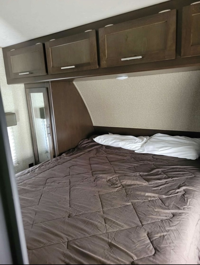 This RV rental bedroom features a bed with a brown comforter, white pillows, and dark wood overhead storage cabinets.. Forest River Wolf Pack 2016