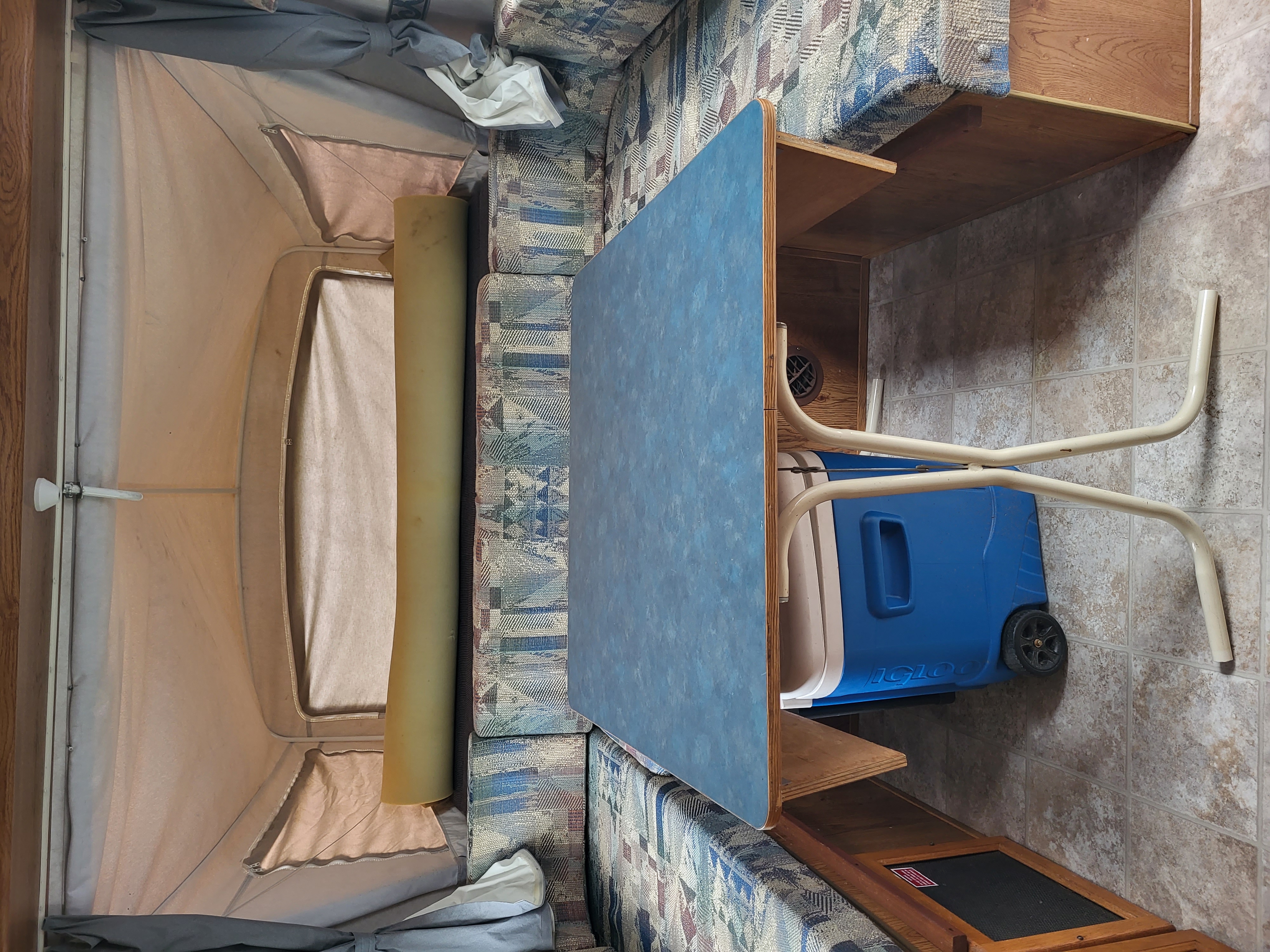 An RV rental interior shows a pop-up camper's fabric sleeping area, cushioned dinette, blue table, and a blue Igloo cooler on the tiled floor.. Aerolite Kodiak 2000