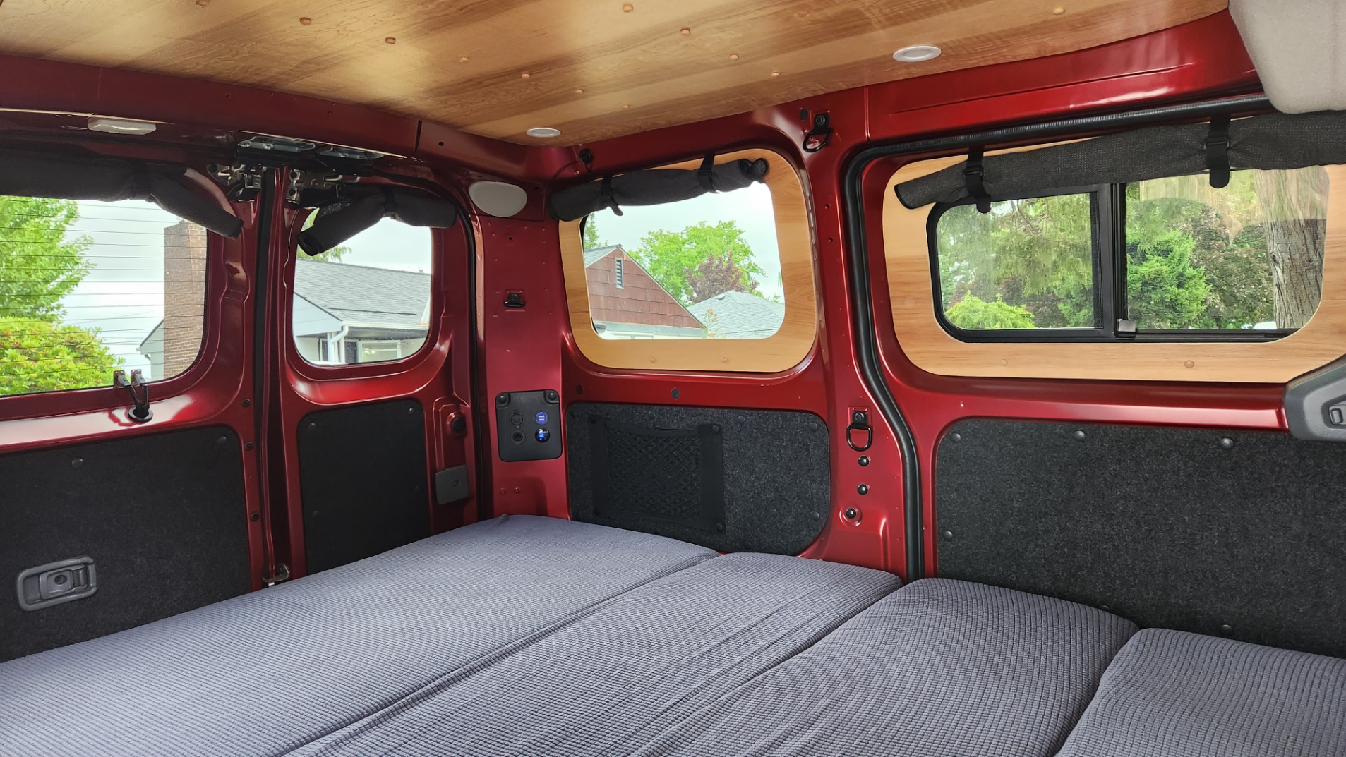 Inside a red camper van ready for RV rental. Features wood ceiling, custom windows, comfy bed & power outlets.. Nissan NV200 SV 2021
