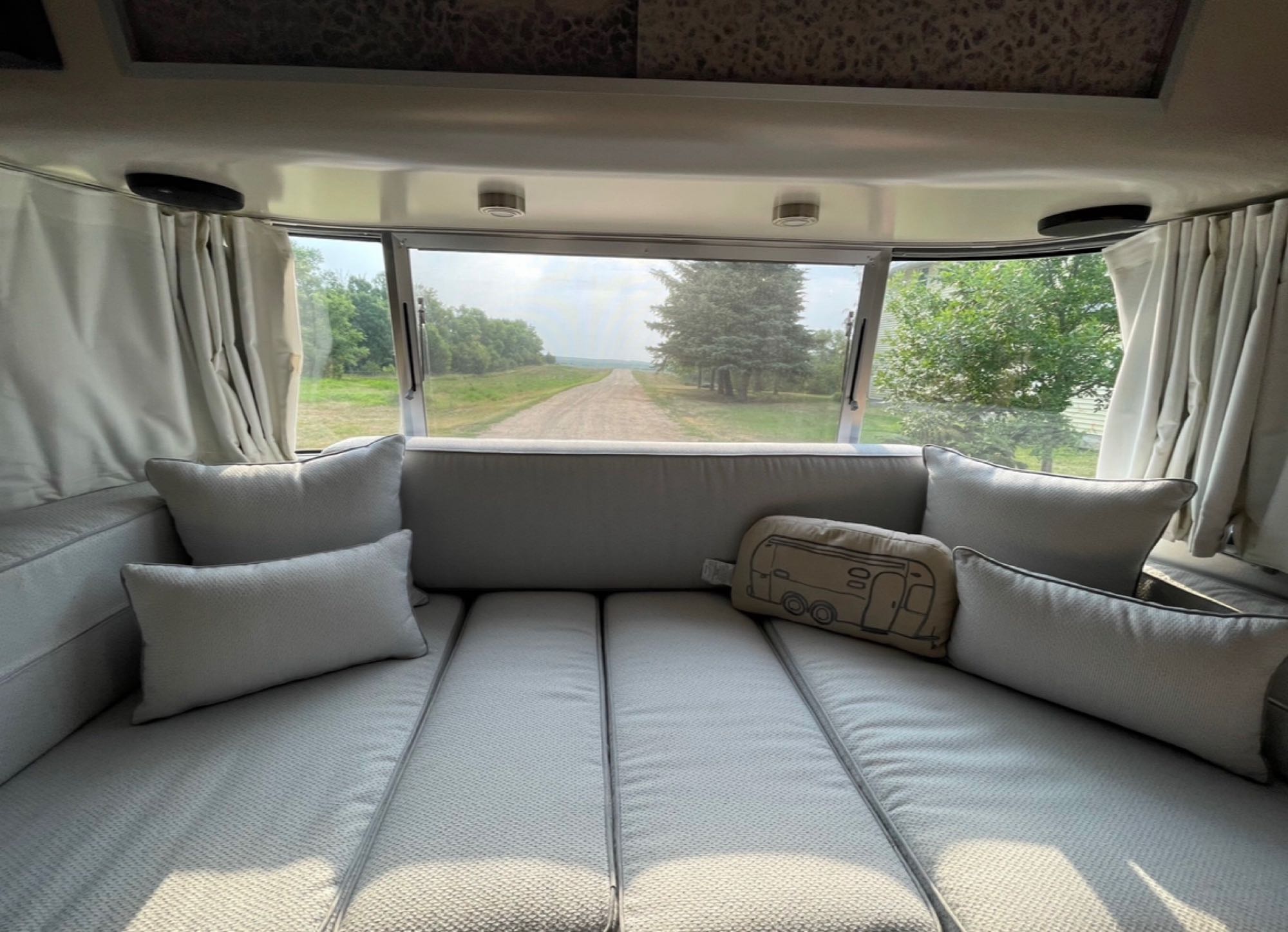 Looking out the back window of an RV rental: a plush bed/lounge area faces a dirt road & trees.. Airstream International 2020