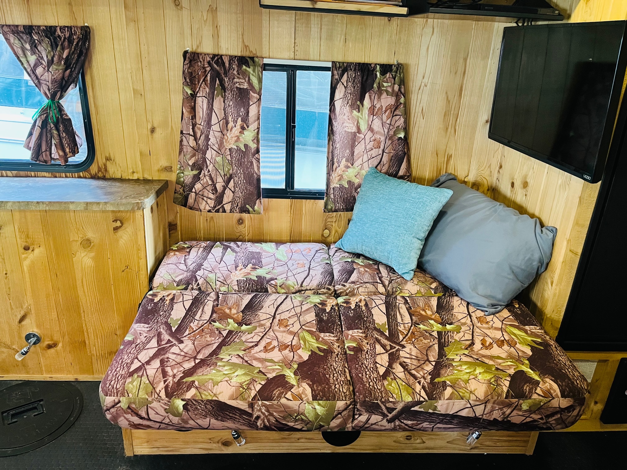 Rustic RV rental interior: wood-paneled walls, camo sofa/bed, multiple windows, and a wall-mounted TV for a cozy stay.. Forest River Cherokee Grey Wolf 2015
