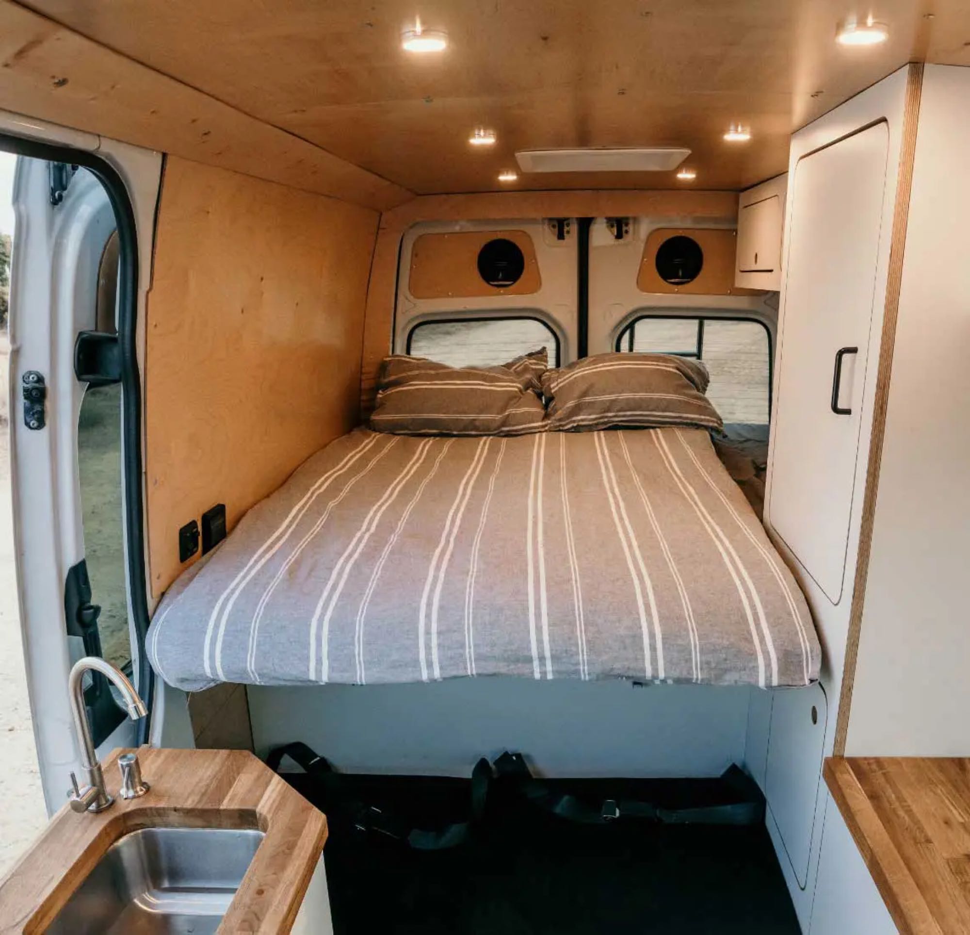 Cozy RV rental camper van interior: large bed with striped linen, compact kitchen sink, and warm wood-paneled walls/ceiling.. Mercedes-Benz Sprinter 2023