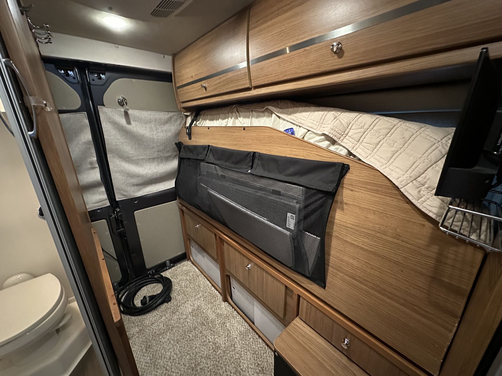 Cozy RV rental interior with wooden cabinets, a bed, and built-in storage. Features a toilet near the entrance.. Winnebago Travato 2017