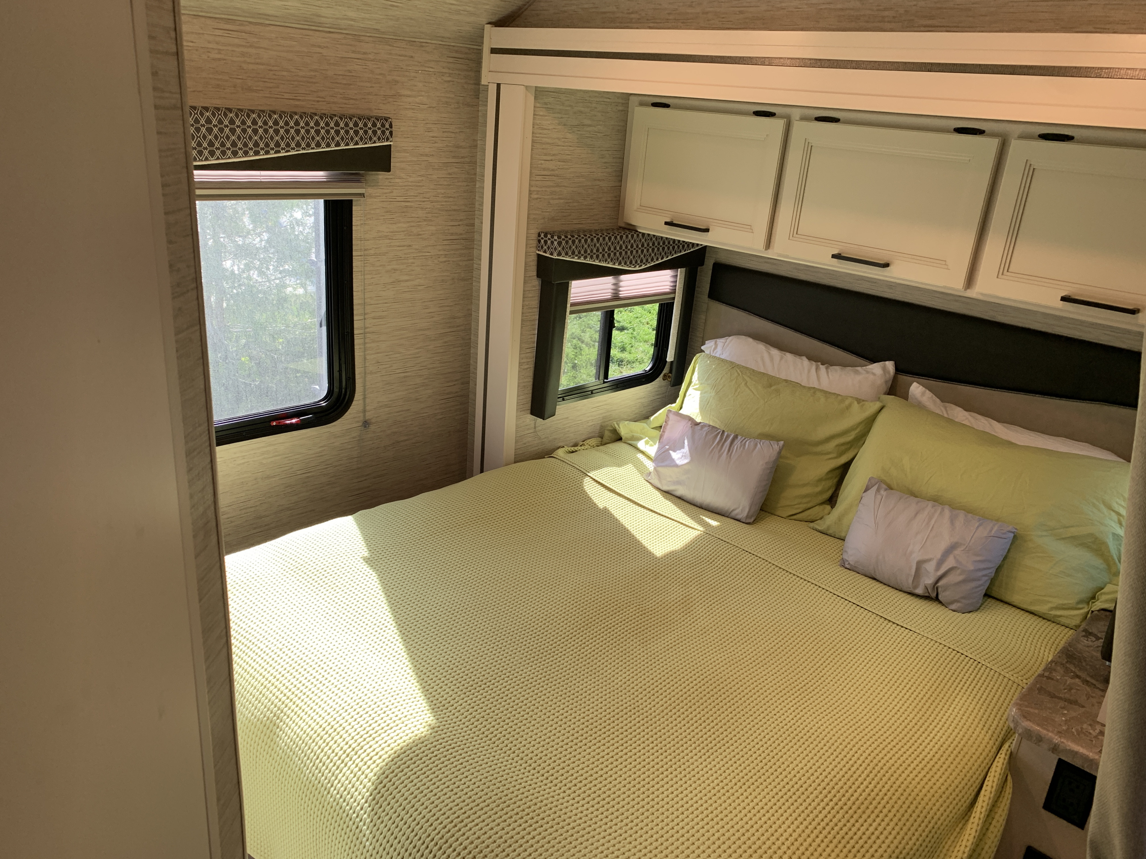 Cozy RV rental bedroom. Features a comfy bed, green blanket, light shining on it, windows, and overhead storage.. Thor Chateau 2023