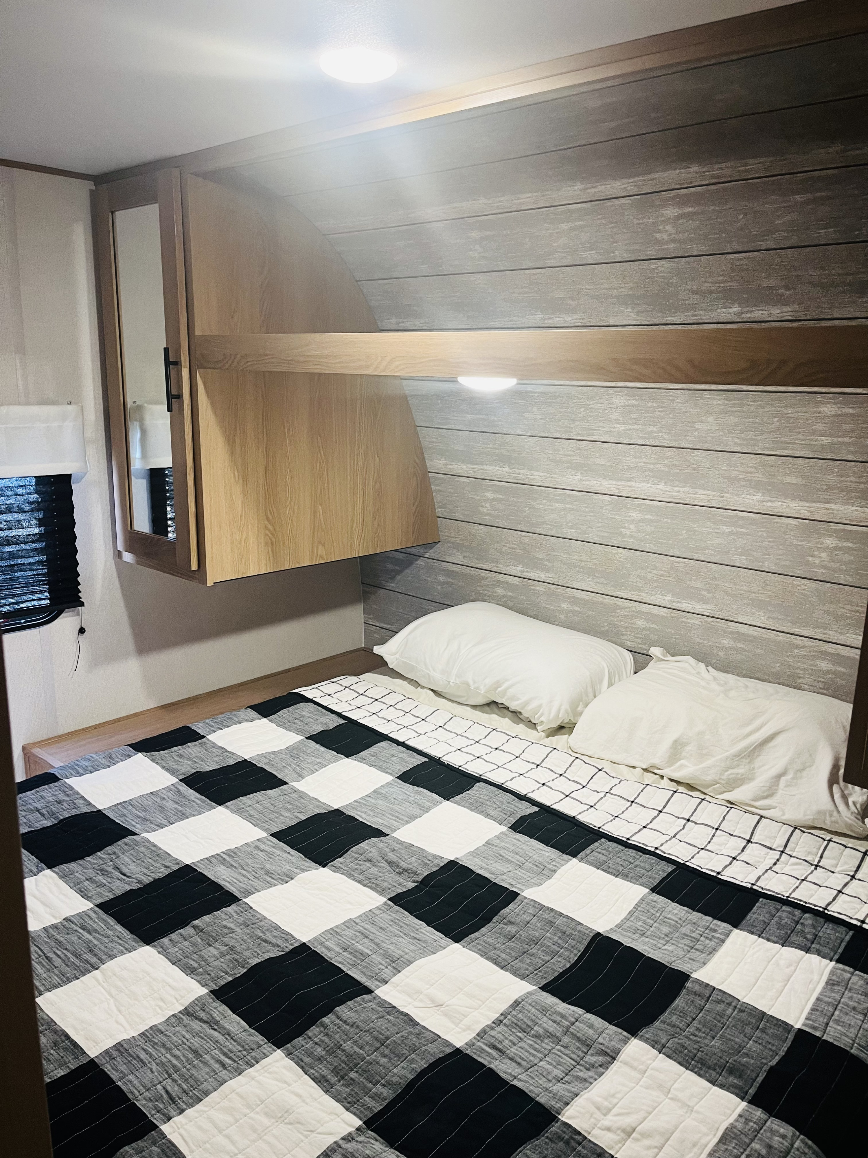 Cozy RV rental bedroom featuring a black and white checkered bedspread, overhead storage, and light wood paneling.. Forest River Prime Time Avenger 24BHSLE 2023