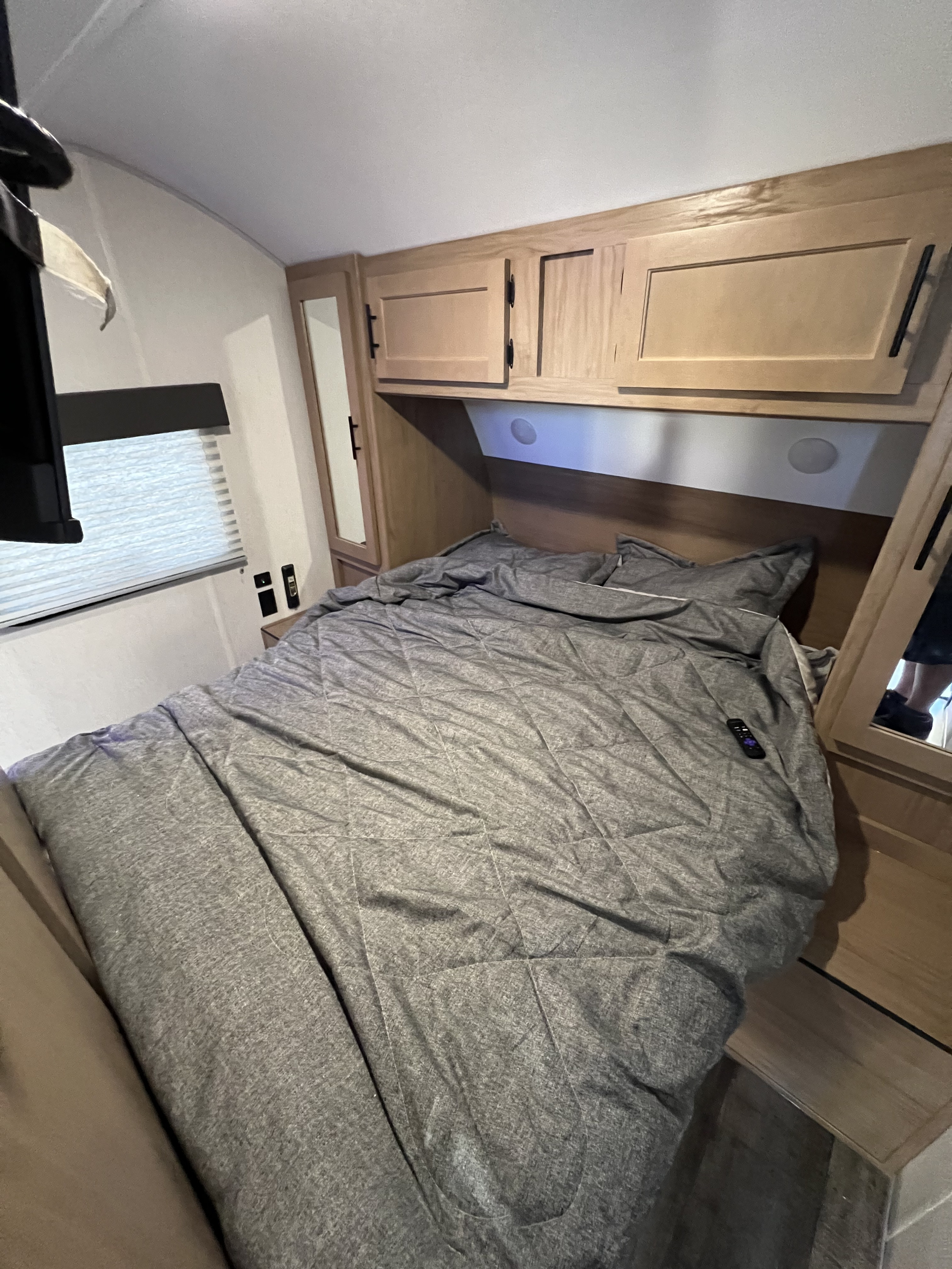Inside an RV rental: a compact bedroom with a grey bed, overhead wood cabinets, a window, and small mirrors.. Forest River Cherokee Grey Wolf 26DBH 2023