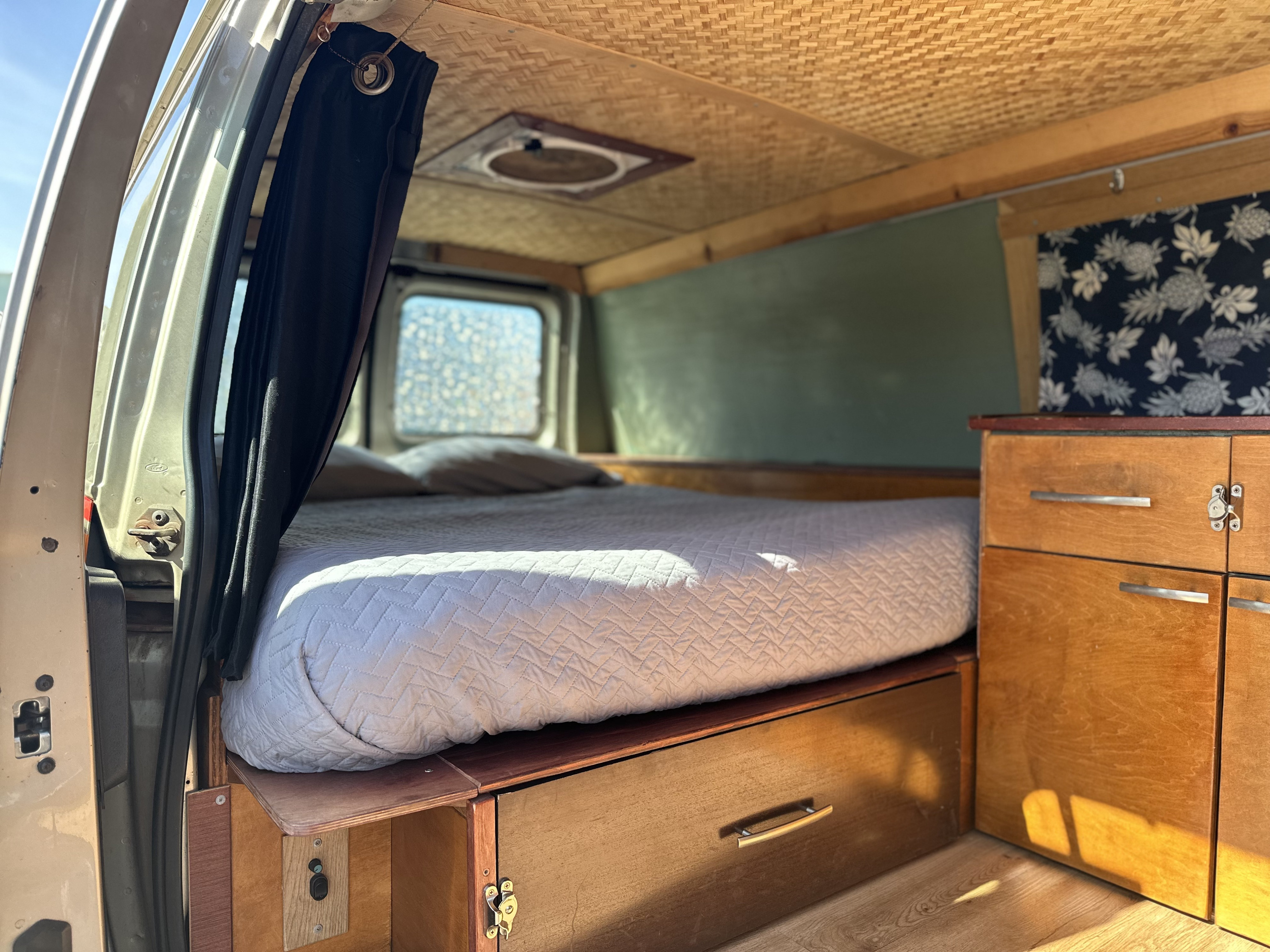 RV rental's cozy interior, showing a full bed with under-bed storage, wooden cabinetry, and a woven bamboo ceiling.. Ford E250 2009