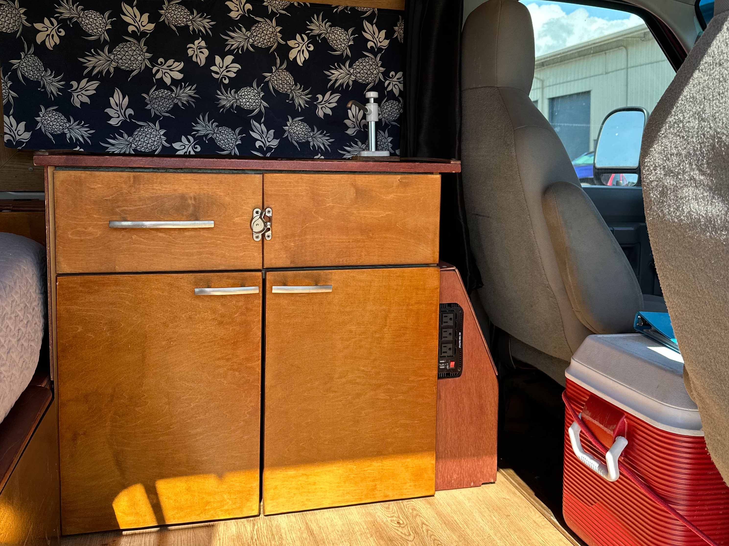 An RV rental van interior featuring custom wooden cabinets, pineapple-patterned fabric, a power inverter, and a red cooler.. Ford E250 2009