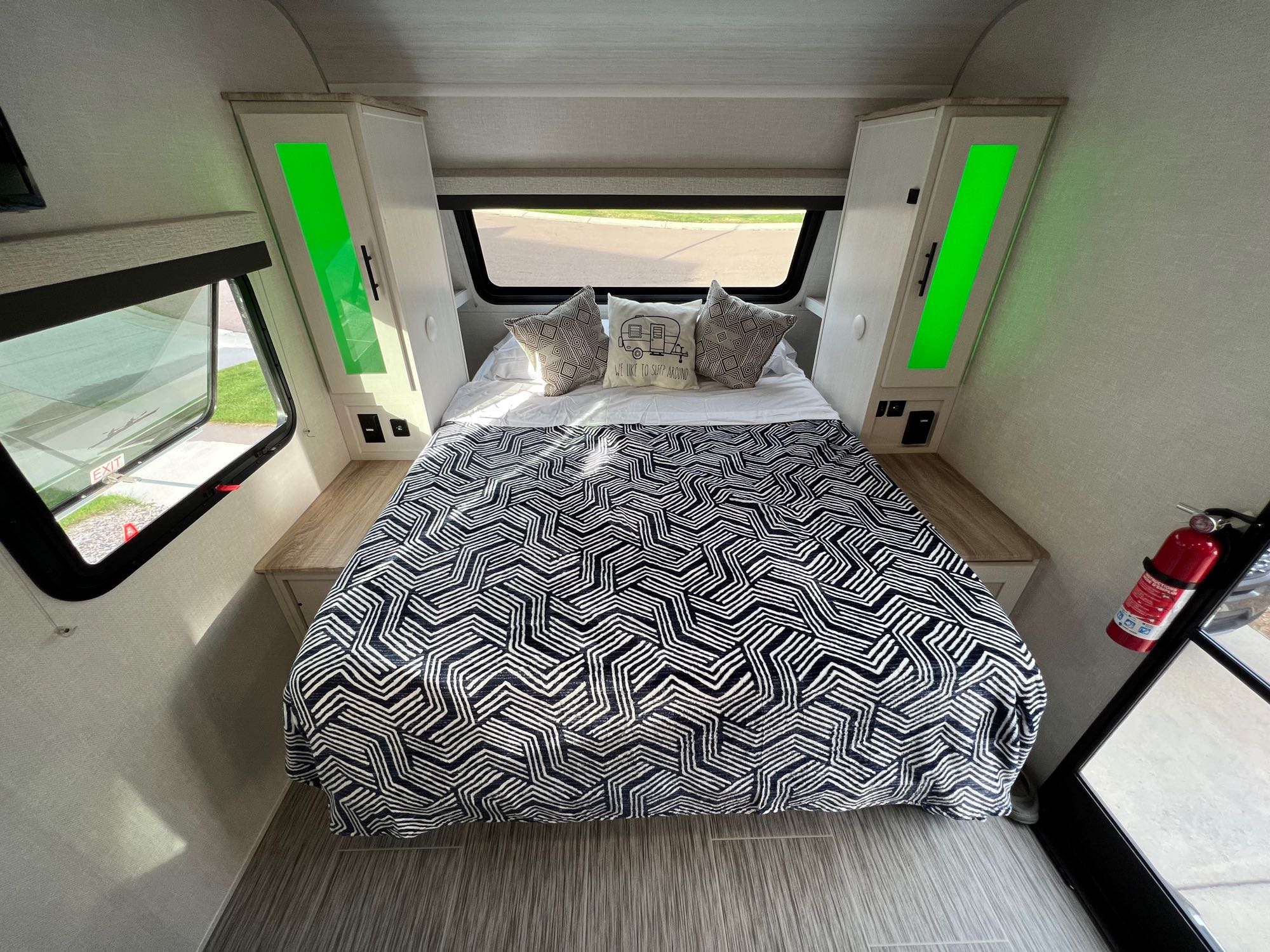 This RV rental bedroom features a large bed with a black/white geometric comforter, "Sleep Around" pillow, green cabinet lights, and an exit window.. No Boundaries 19.3 2023