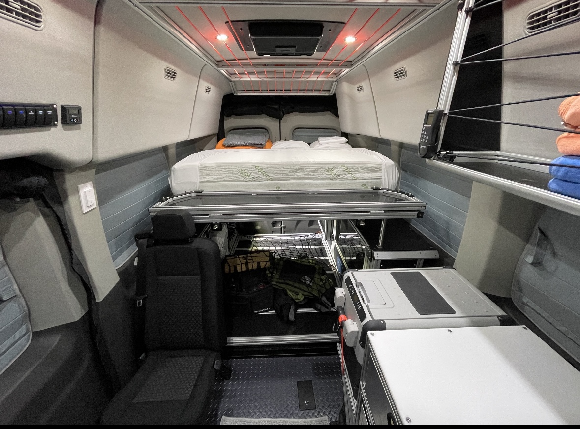 Compact RV rental interior featuring a raised bed, under-bed storage, and portable fridge for travel comfort.. VanDoit Liv AWD Camper Van 2022