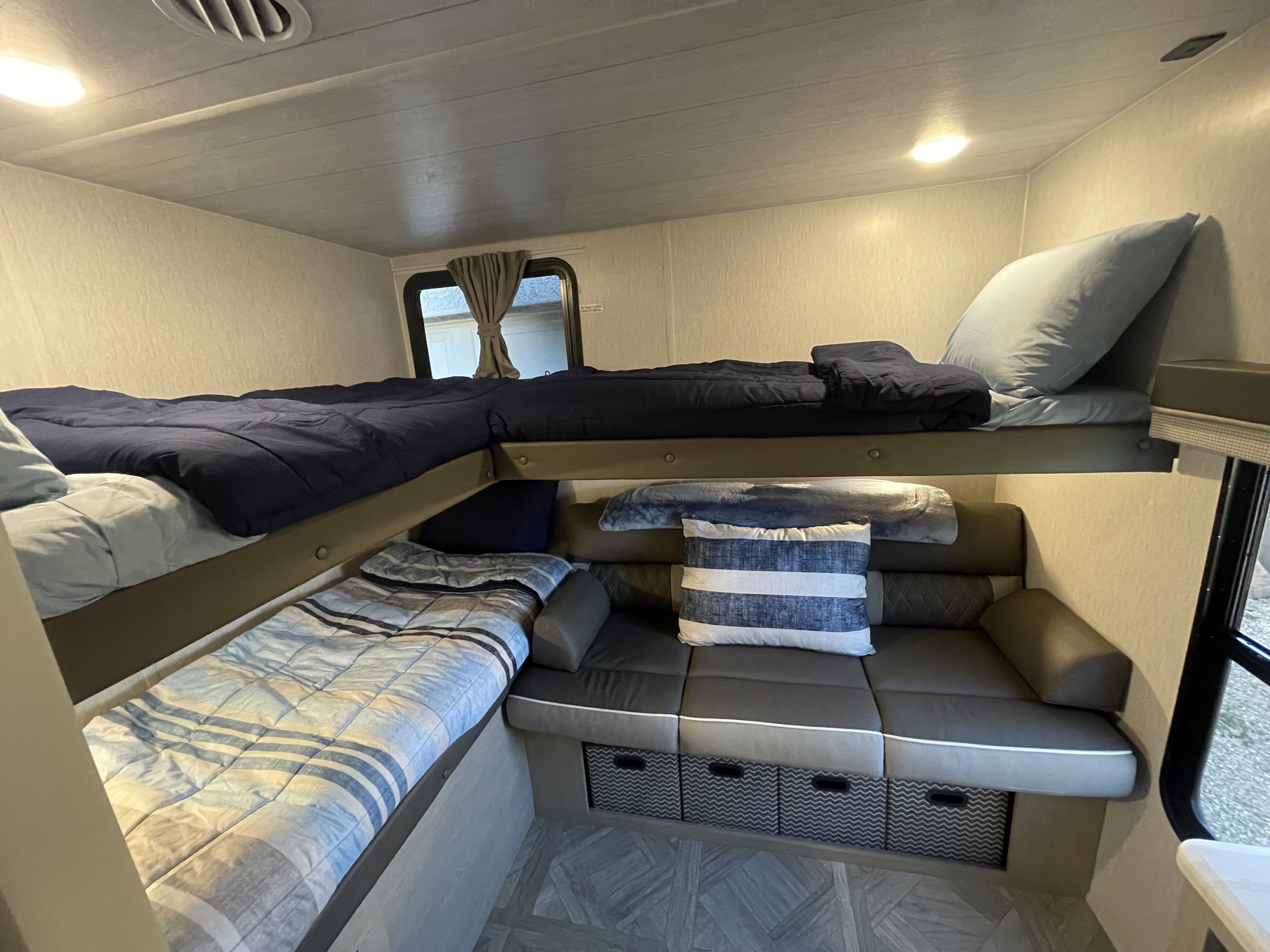 RV rental's compact interior: features cozy bunk beds (top dark blue, bottom striped) and a gray sofa with storage for travelers.. Forest River Wildwood X-Lite 2023
