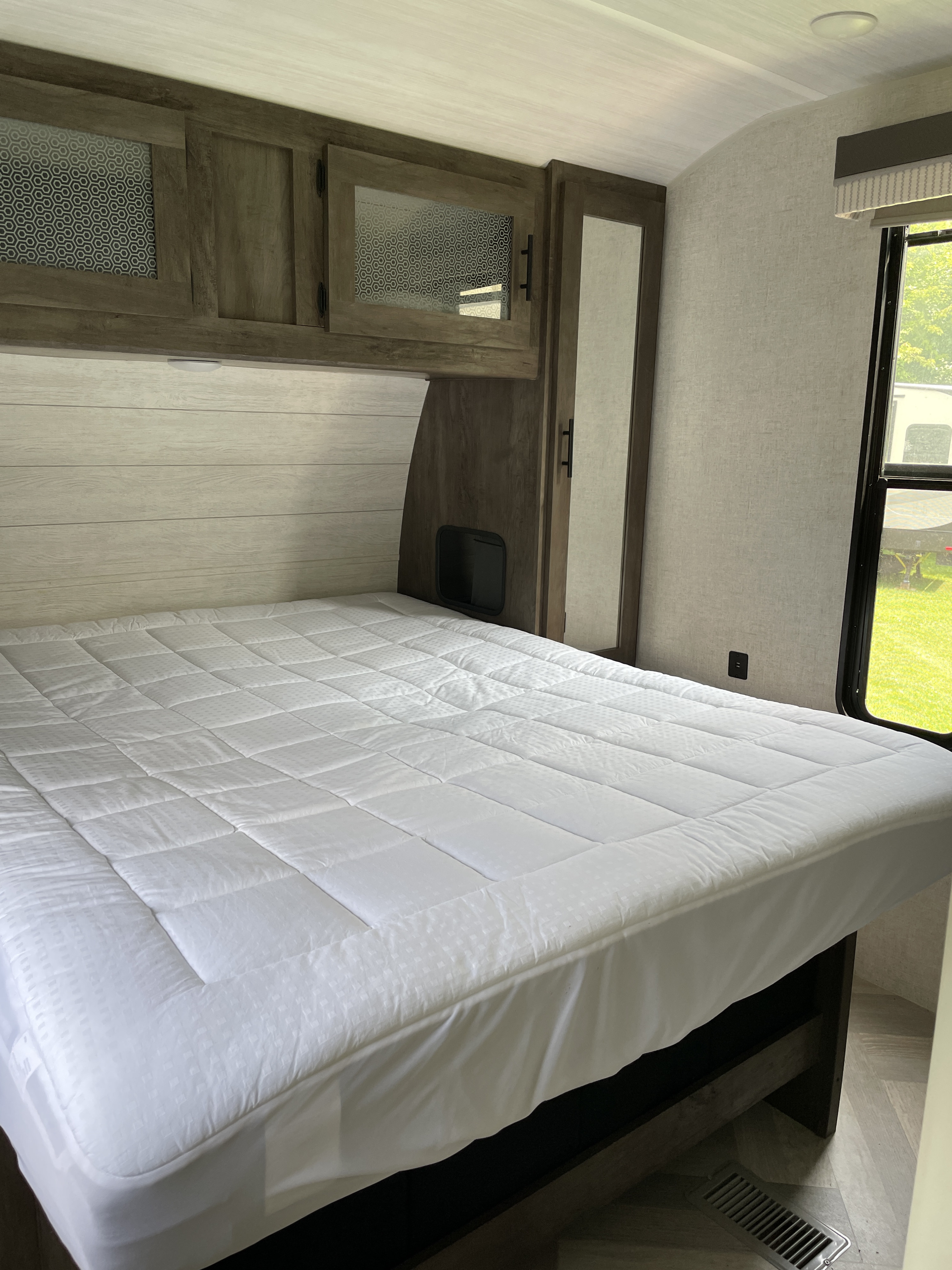 Inside an RV rental bedroom, featuring a white bed, overhead cabinets with patterned glass, and a window.. Forest River Wildwood 2021